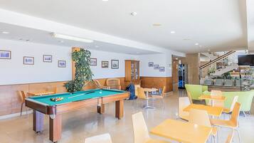 A pool table with chairs, a dining area with tables and chairs, framed pictures on the wall, and a staircase in the background.