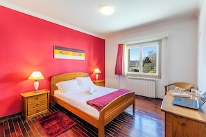 Quarto com cama de madeira, duas mesas de cabeceira com luminárias, um tapete vermelho, uma janela com vista e uma escrivaninha com um laptop.