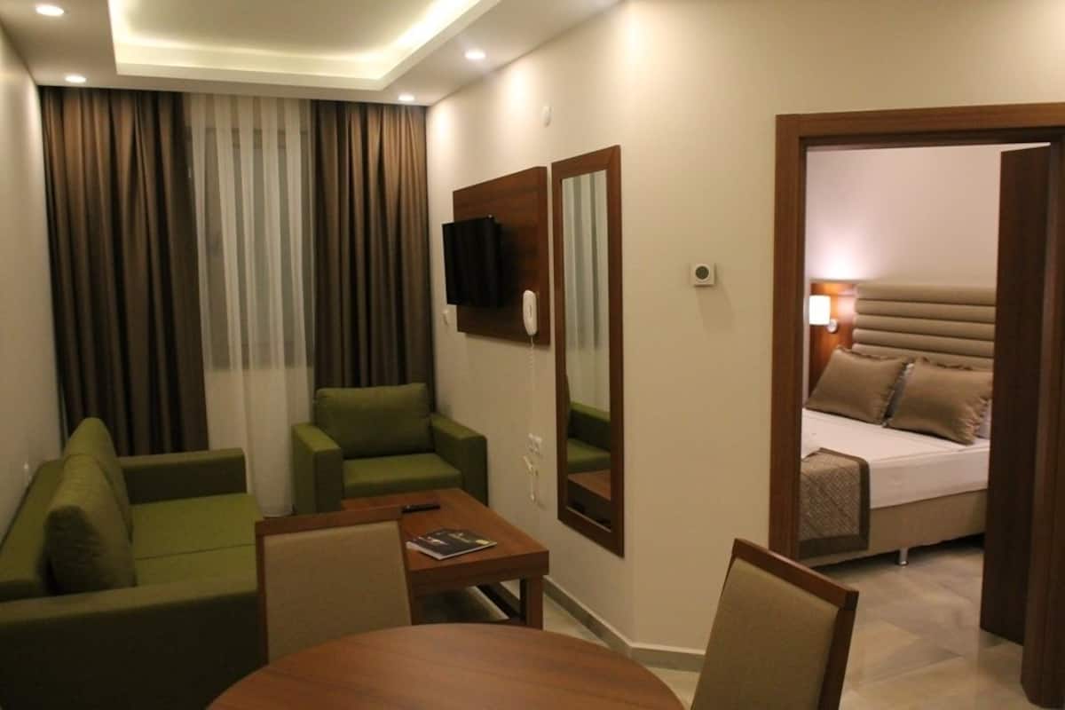 Habitación de hotel con sofá verde, una mesita, televisor montado en la pared y cama con ropa de cama color beige.