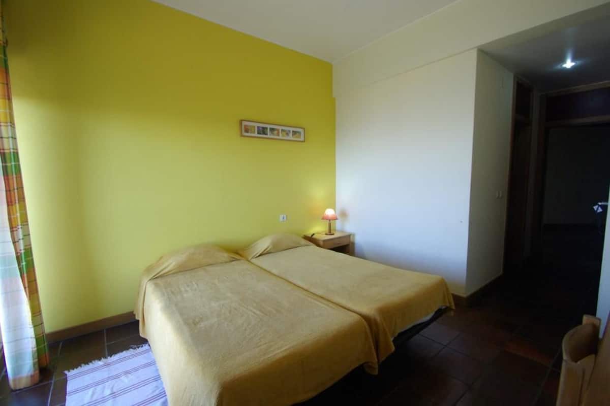 Ein Zimmer mit zwei Betten, einem Nachttisch mit Lampe, einem Wandbild und einer Tür zu einem anderen Raum.