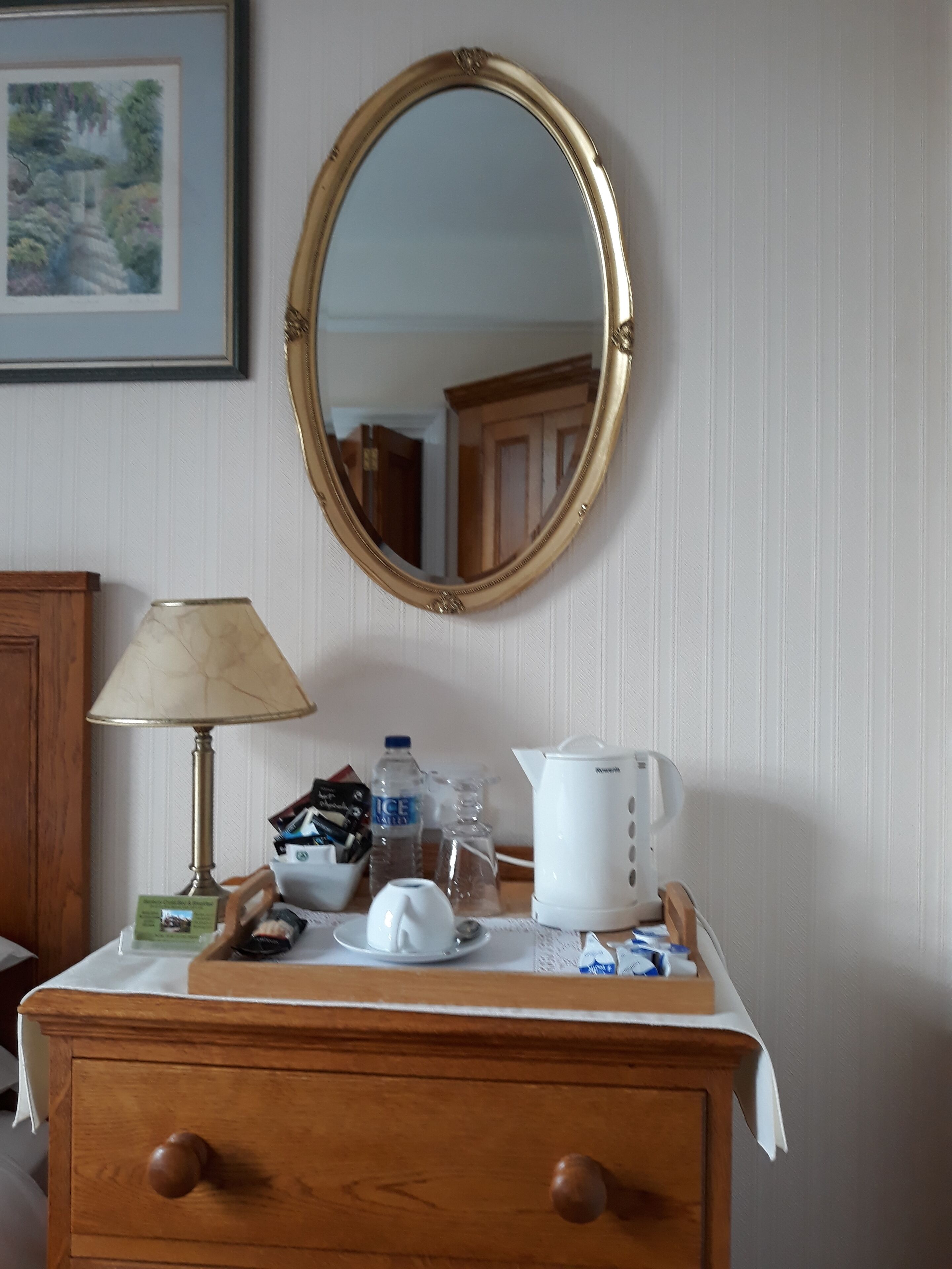 A wooden dresser with a white tablecloth, a lamp, a kettle, a water bottle, and a framed picture on the wall.
