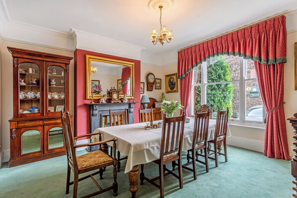 A dining room with a table set for six, surrounded by wooden chairs. A wooden cabinet with glass doors is to the left, and a large mirror hangs above the table.