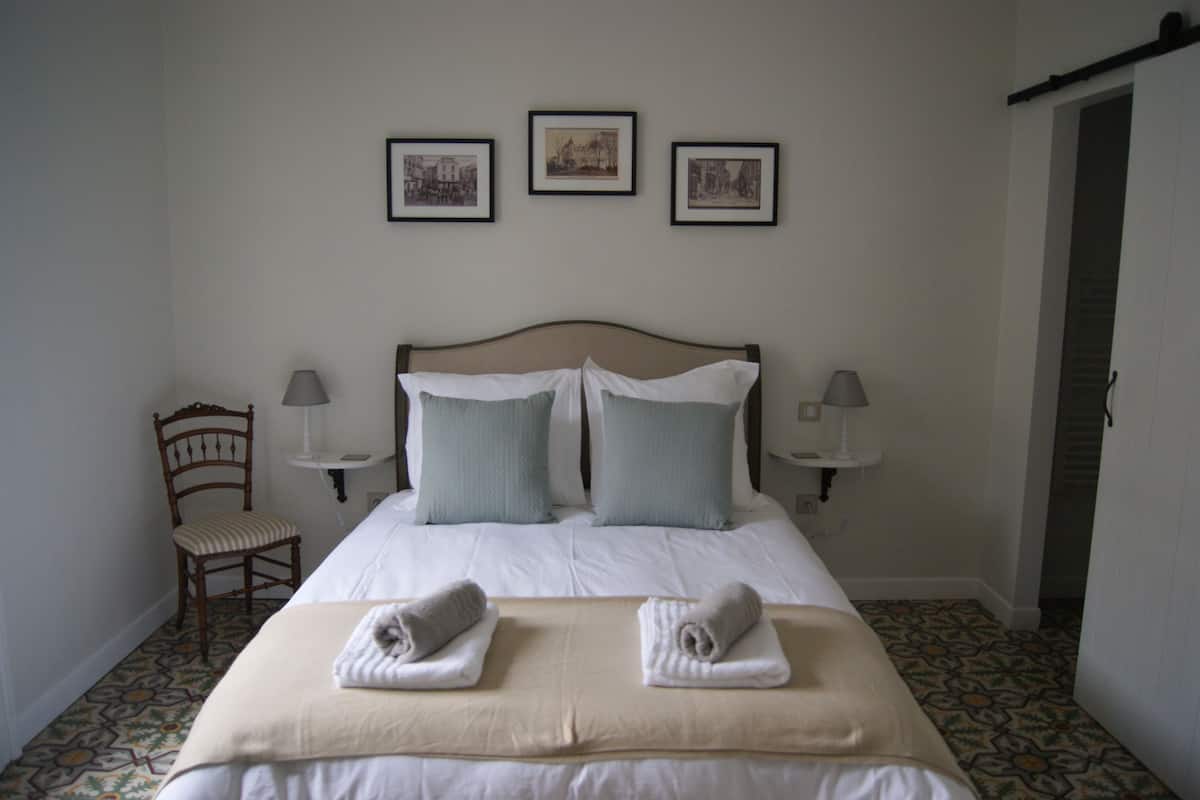 Ein ordentlich bezogenes Bett mit zwei Kissen, ein Sessel und drei gerahmte Bilder an der Wand.