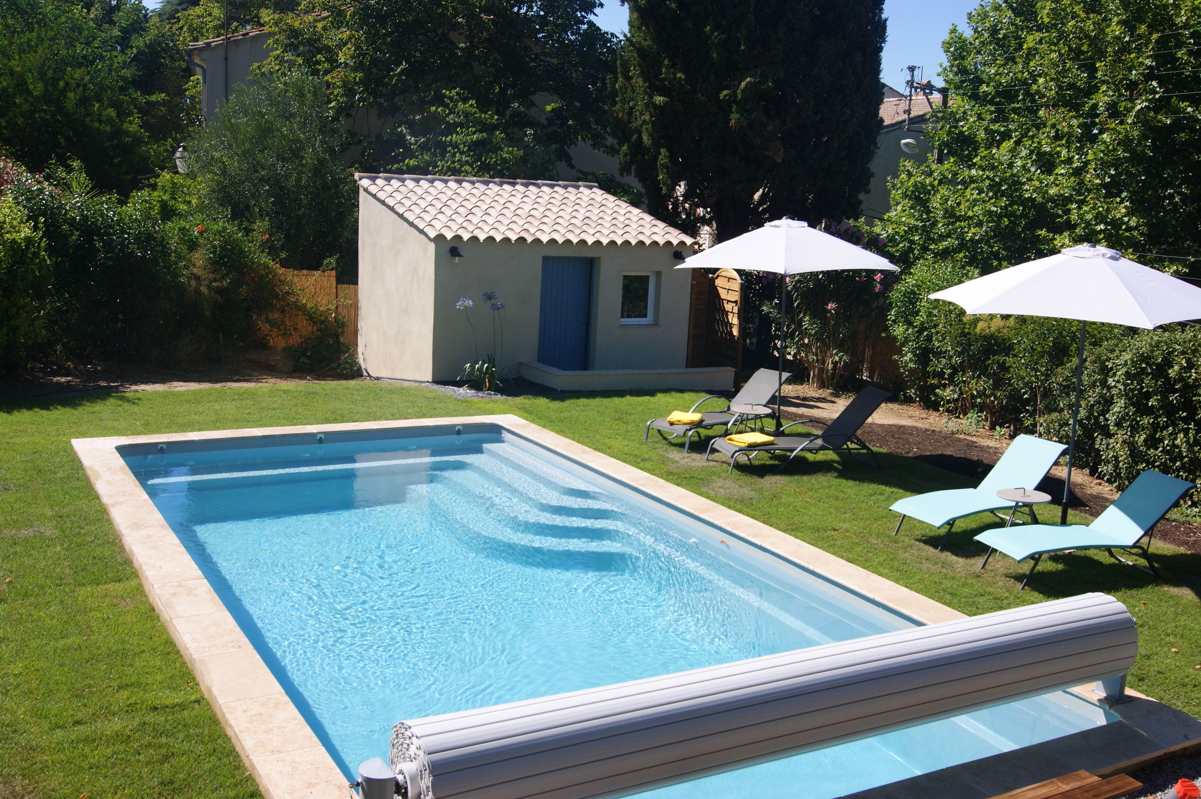 A rectangular swimming pool with a tiled deck, surrounded by a well-maintained lawn. There are two lounge chairs and two umbrellas providing shade.