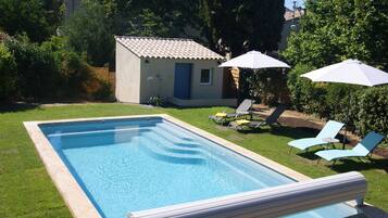 A rectangular swimming pool with a tiled deck, surrounded by a well-maintained lawn. There are two lounge chairs and two umbrellas providing shade.