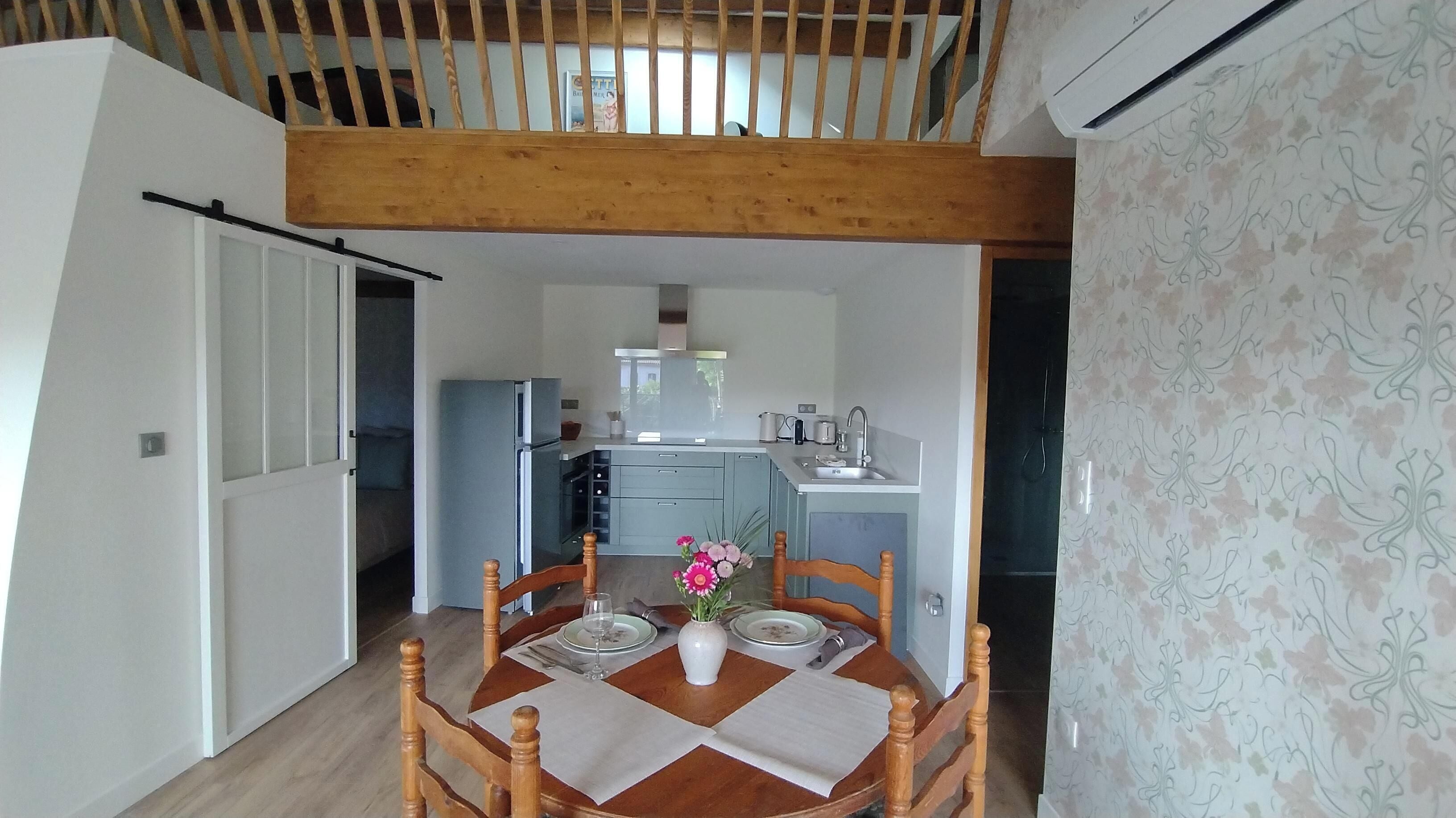 Un comedor con una mesa de madera, sillas y un jarrón de flores. Se ve una cocina al fondo con electrodomésticos modernos.