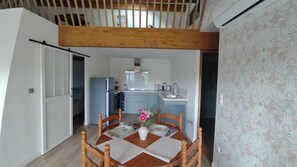 A dining area with a wooden table, chairs, and a vase of flowers. A kitchen is visible in the background with modern appliances. - Le Petit Molière Chambres d'Hôtes  (Servian)