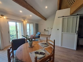 Un comedor con una mesa de madera dispuesta para dos personas, un jarrón con flores y vistas al balcón a través de puertas correderas abiertas.
