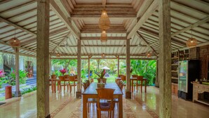 Un espace repas abrité, avec une longue table et des chaises, entouré de piliers en bois et agrémenté de luminaires suspendus.