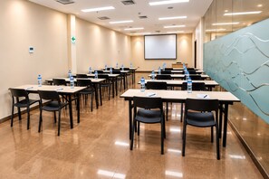 A conference room with tables, chairs, water bottles, a projector screen, and a wall with a wavy design.