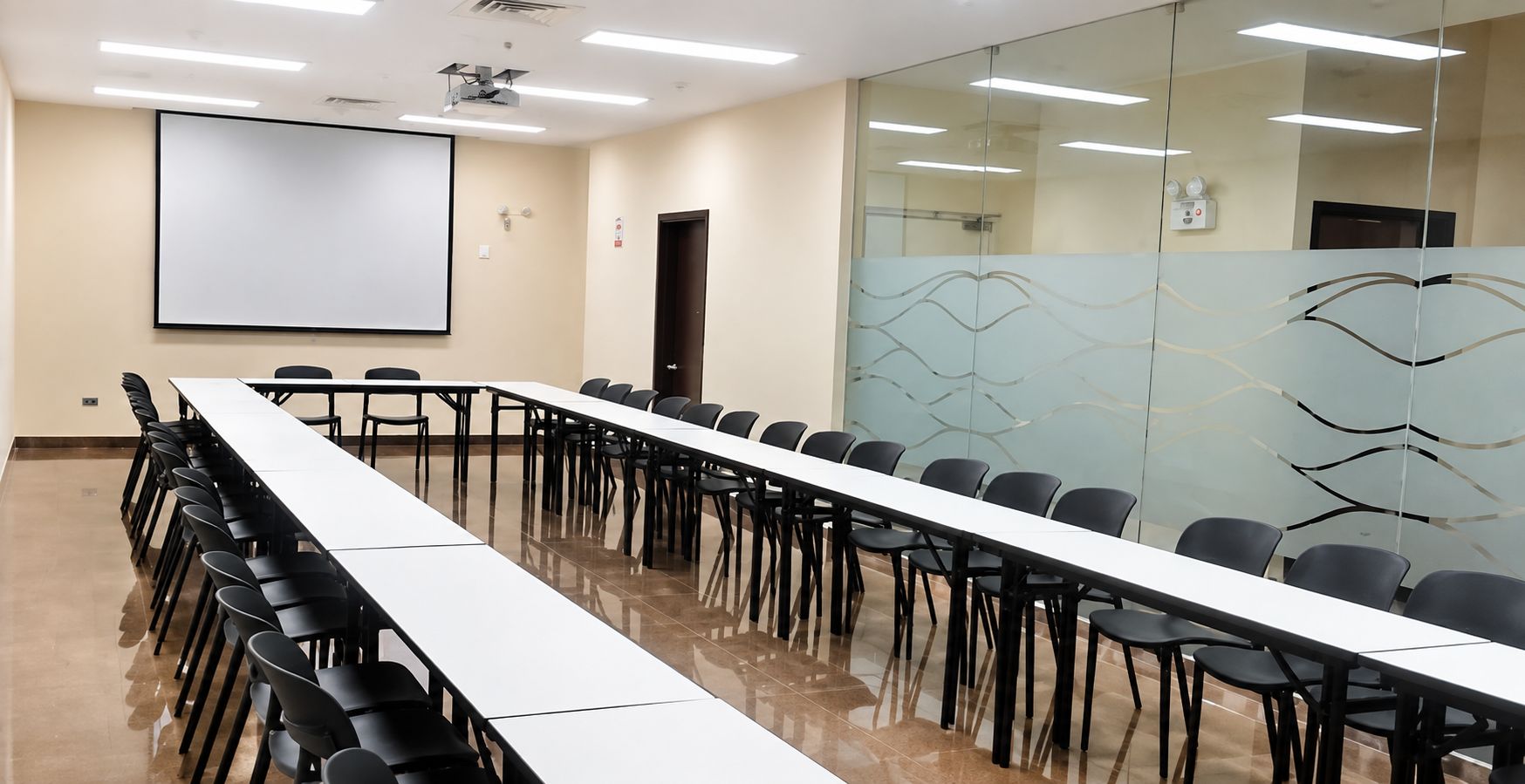 A conference room with a long table, chairs, a projector screen, and a glass partition with a wavy design.