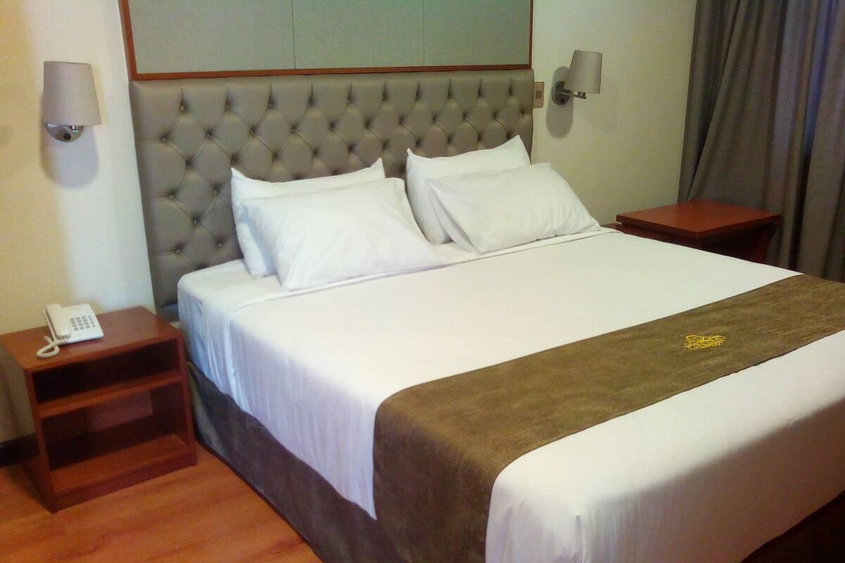 A hotel room with a large bed, a wooden nightstand with a phone, and a headboard with wall sconces.