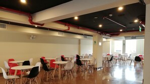Un espacio interior amplio con varias mesas y sillas, tuberías rojas en el techo y grandes ventanales que permiten la entrada de luz natural.