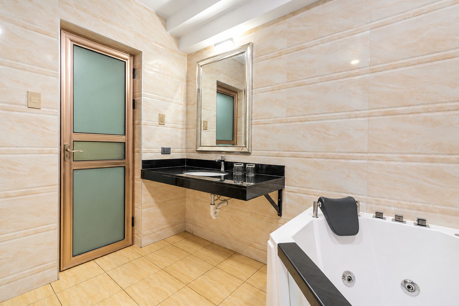 A bathroom with a marble wall, a sink with a black countertop, a mirror, a bathtub, and a door with frosted glass panels.