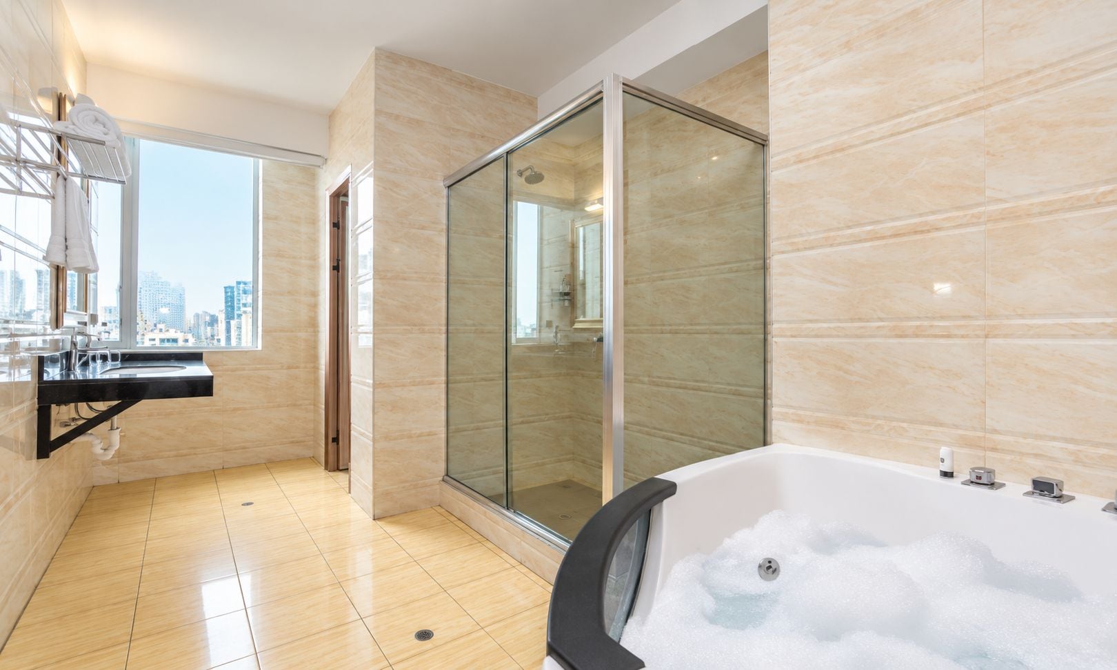 A modern bathroom with a glass-enclosed shower, a large bathtub filled with bubbles, and a city view through the window.