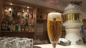 A bar area with a tall glass of beer, a tray of glasses, and a shelf of liquor bottles. - Pension Nadia (Adorf)
