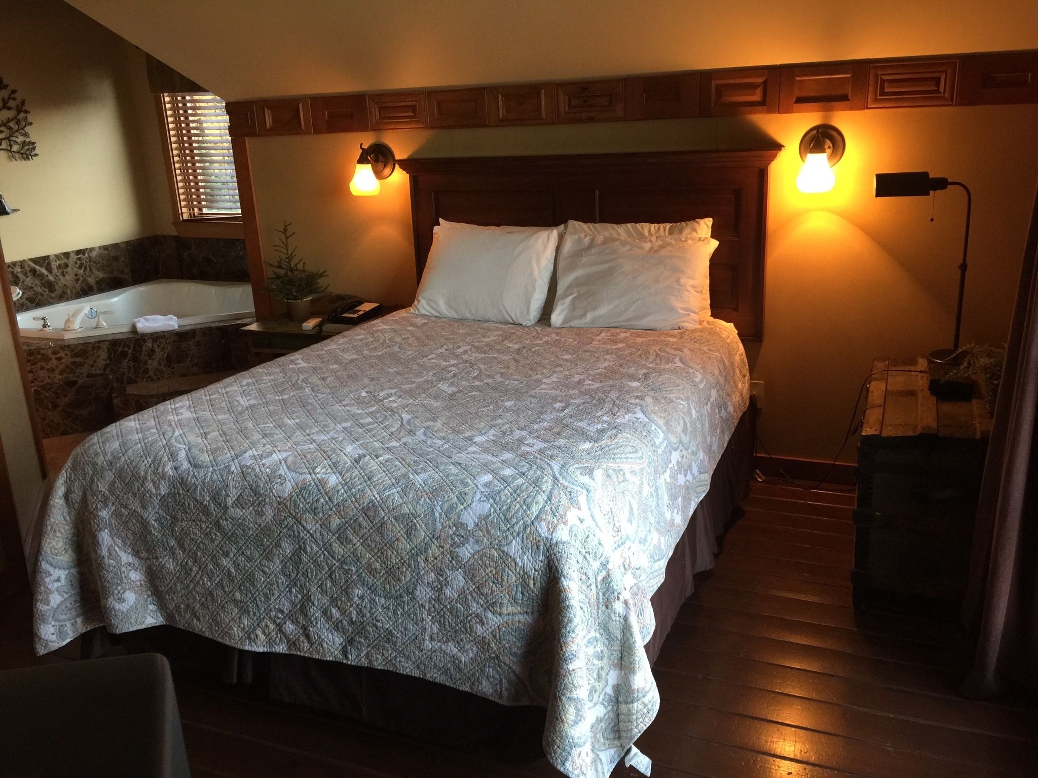 A bedroom with a large bed, a wooden headboard, two wall-mounted lamps, a nightstand, a mirror, and a bathtub.