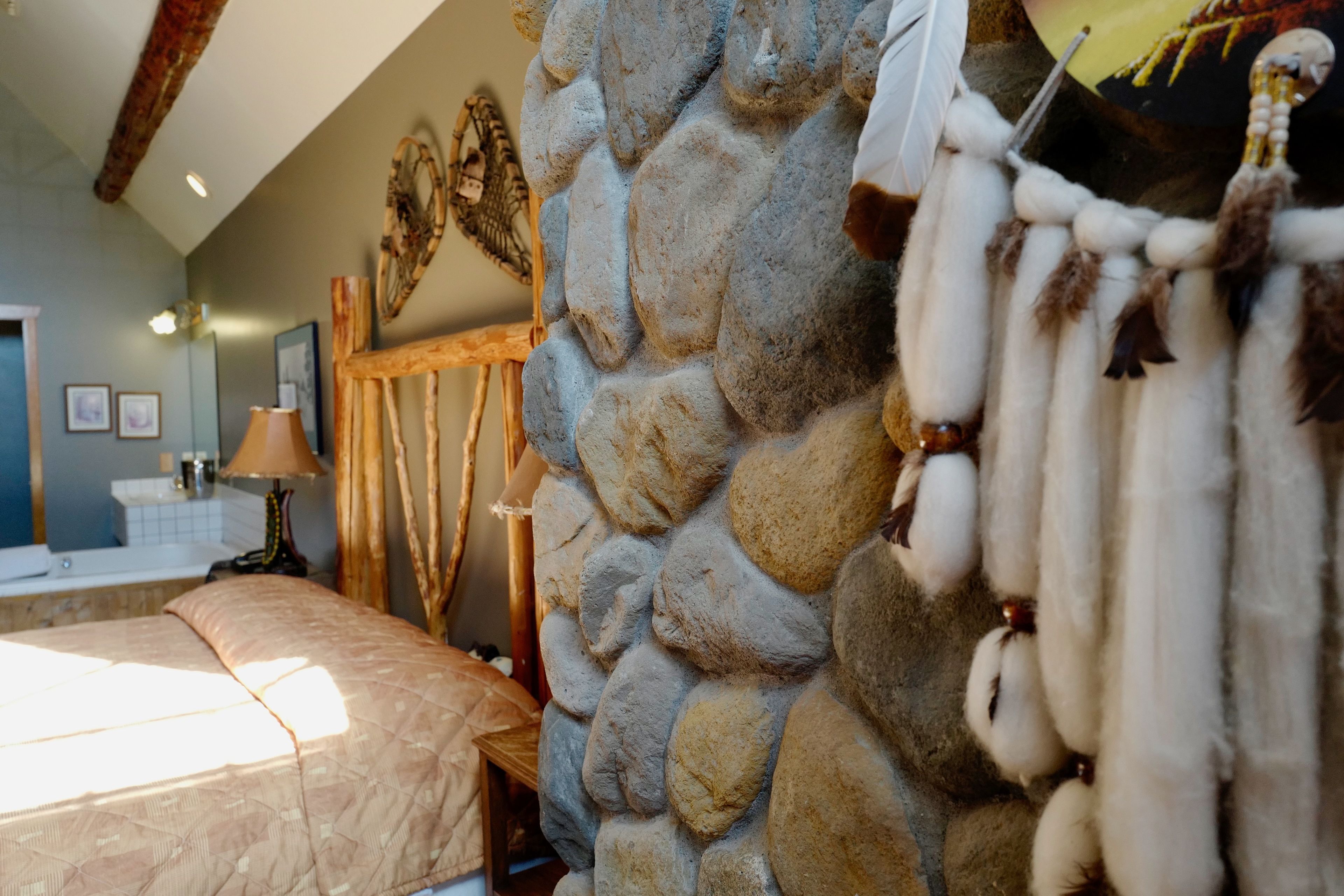 Ein Zimmer mit einer Steinwand, einem Bett mit beiger Bettdecke, einem Holzstuhl, einer Lampe und einem an der Wand hängenden Traumfänger.