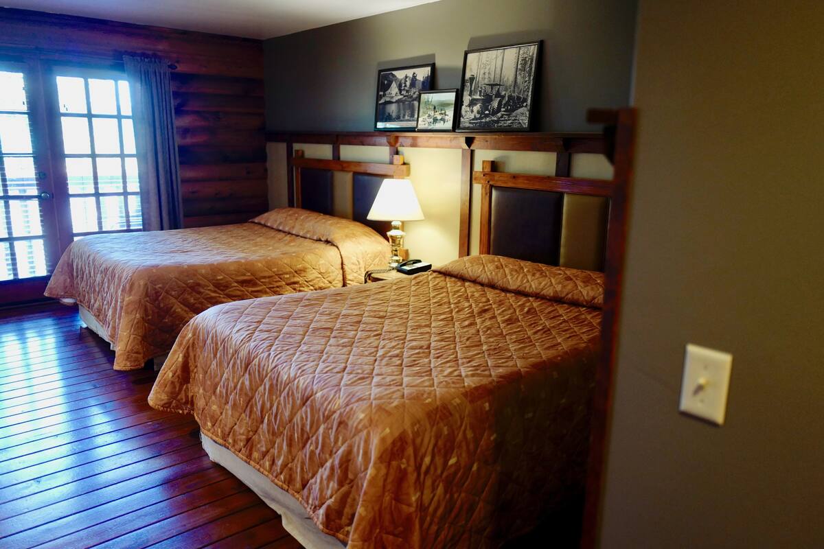 A room with two beds, wooden floors, a lamp on the nightstand, and framed pictures on the wall.