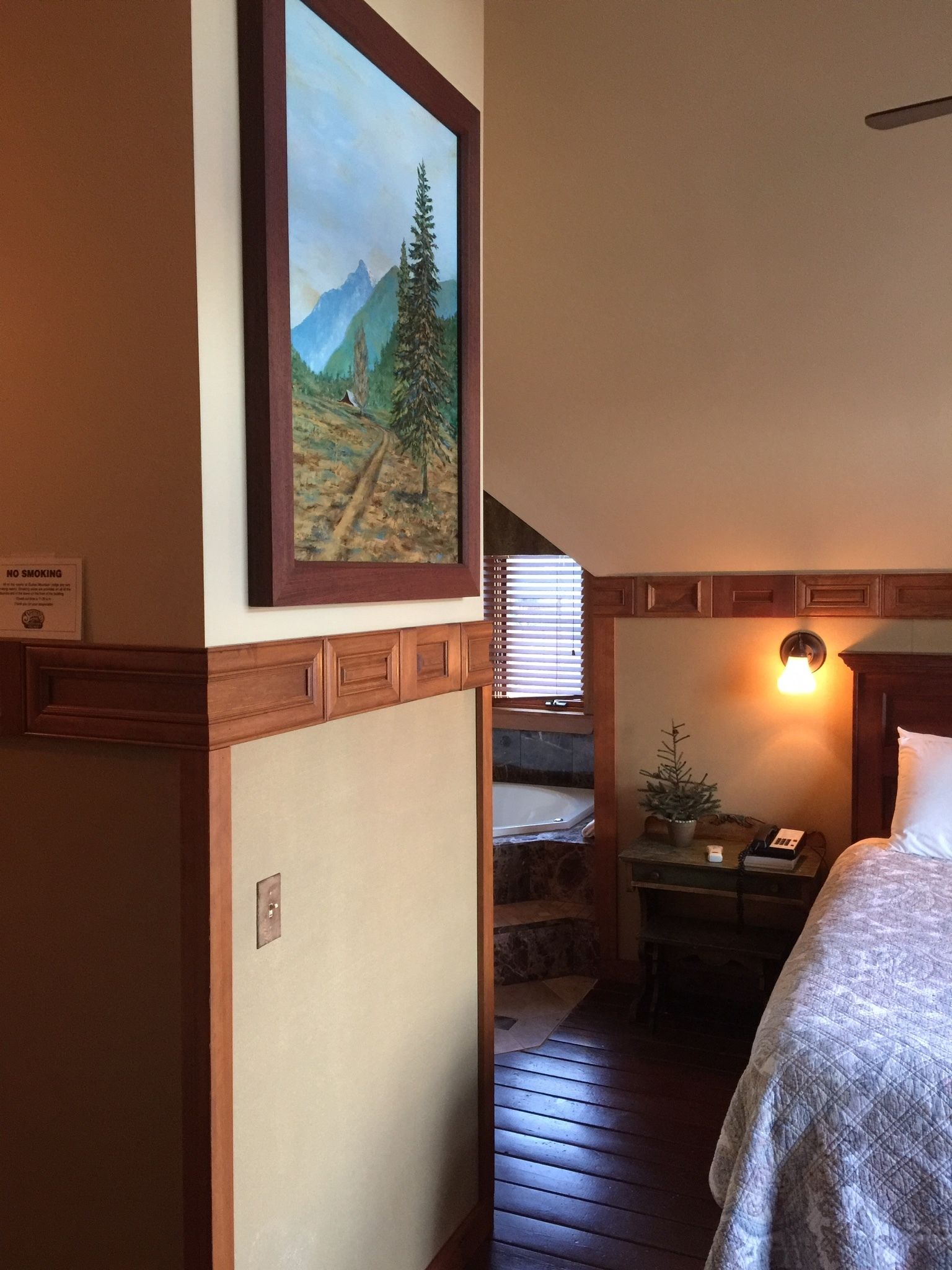 A bedroom with a bed, a nightstand, a lamp, a small table with a plant, and a painting of a mountain landscape.