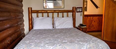 A wooden bed with a patterned bedspread, a framed picture on the wall, and a wooden door with a hanging saw blade.