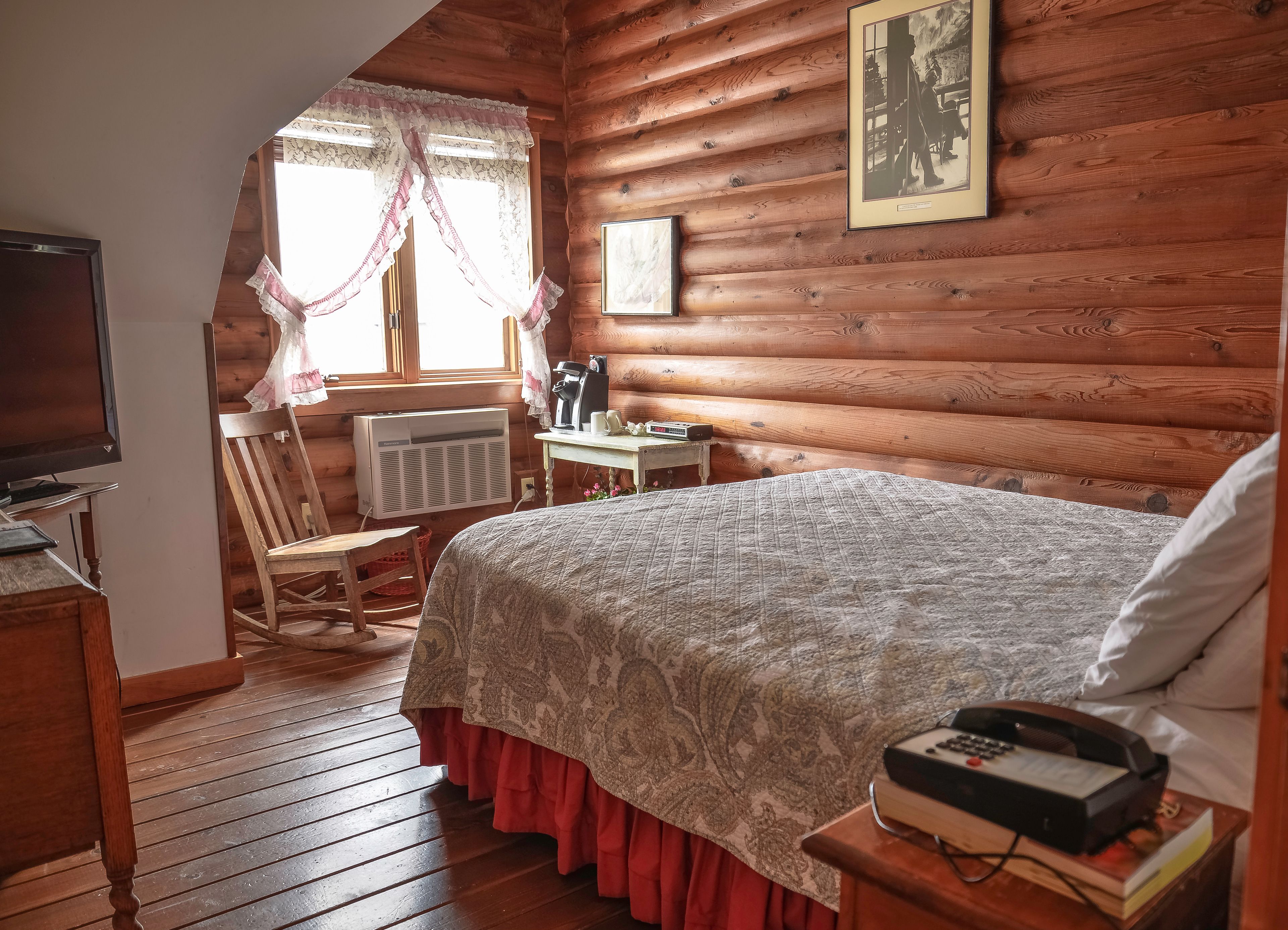 A cozy log cabin bedroom with a bed, a chair, a table with a coffee maker, and a television.