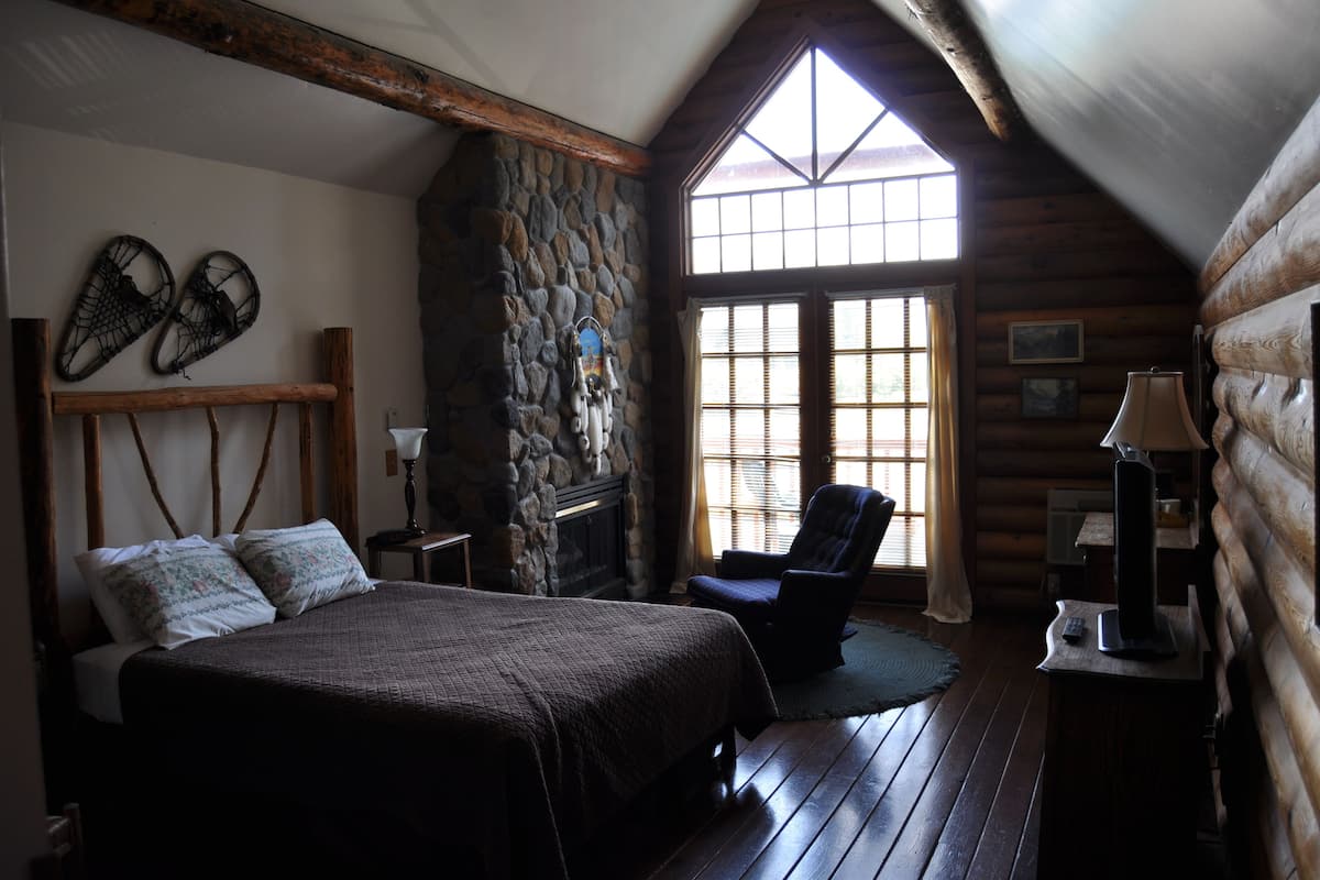 A cozy log cabin bedroom with a large bed, a stone fireplace, a rocking chair, and a window with blinds.