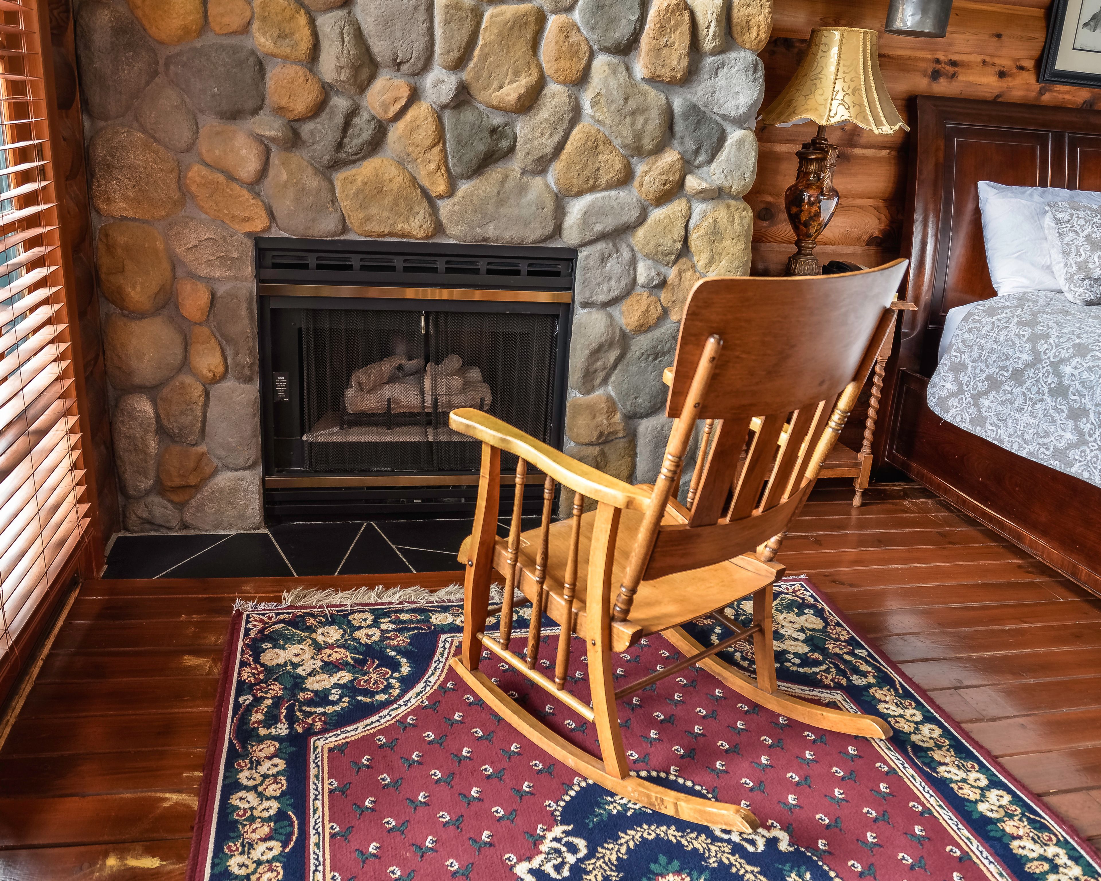 A wooden rocking chair in front of a stone fireplace, a bed with a patterned bedspread, and a wooden floor.