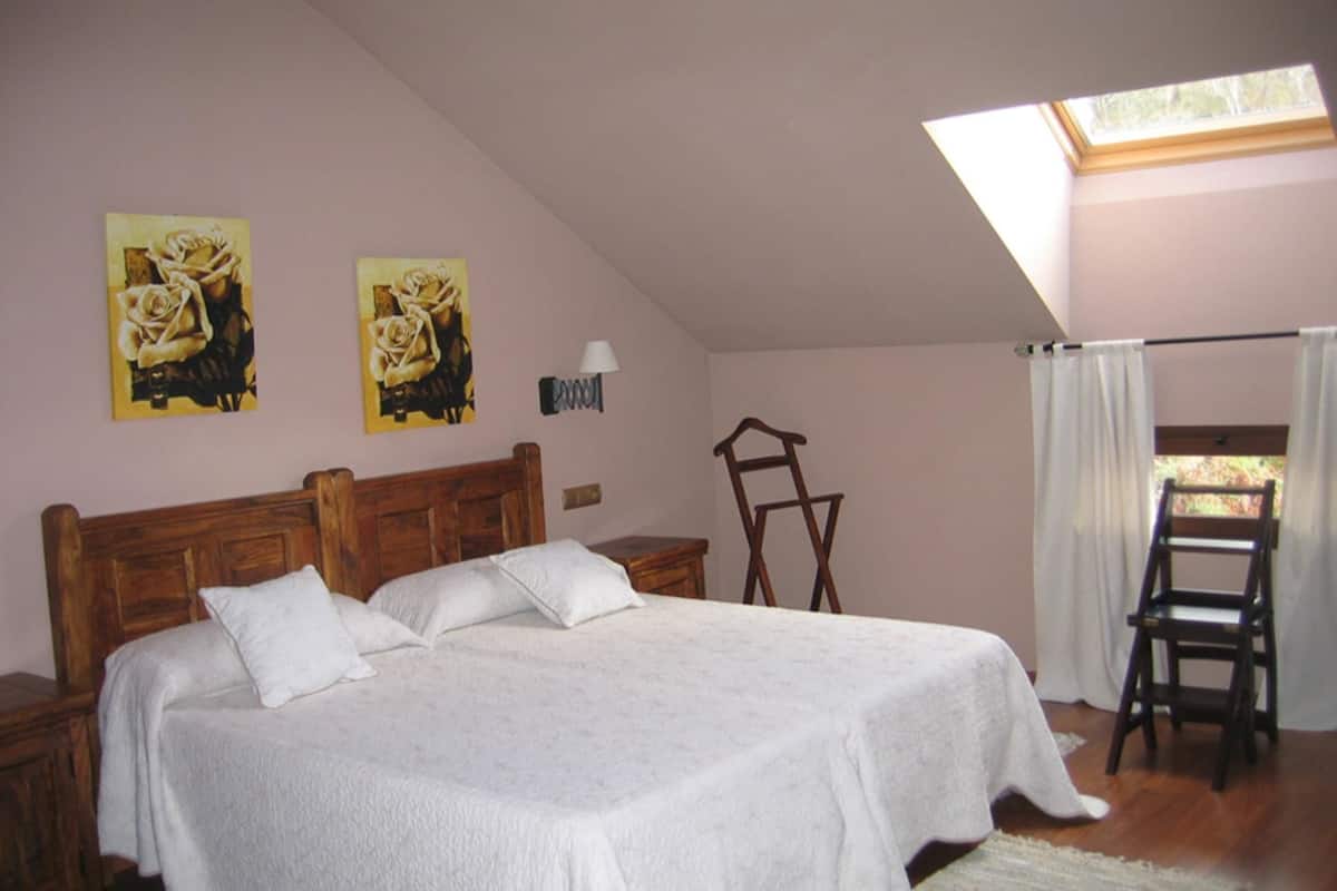 Une chambre à coucher avec un grand lit, une tête de lit en bois, deux tableaux au mur, une table de chevet, une lampe, une chaise en bois et une fenêtre avec des rideaux.