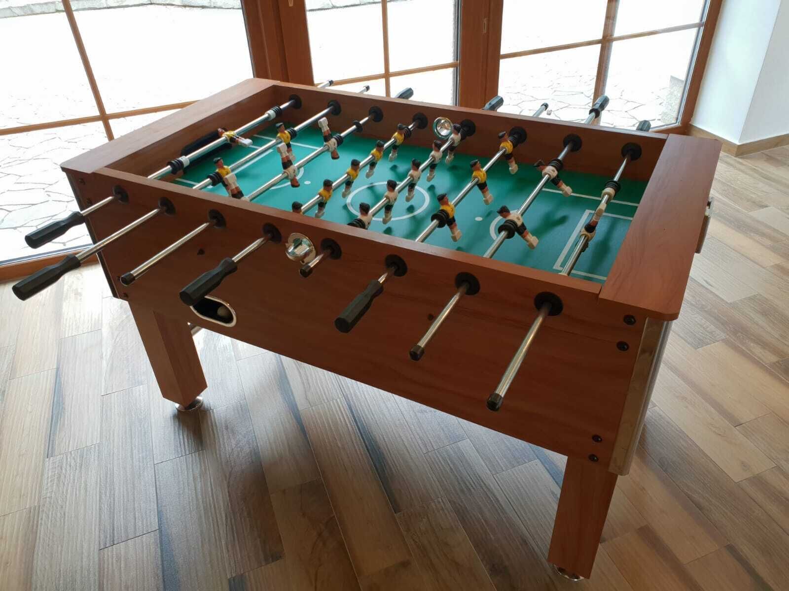 Uma mesa de futebol de madeira com várias barras e peças, sobre um campo de jogo verde, em um quarto com janelas amplas e piso de madeira.