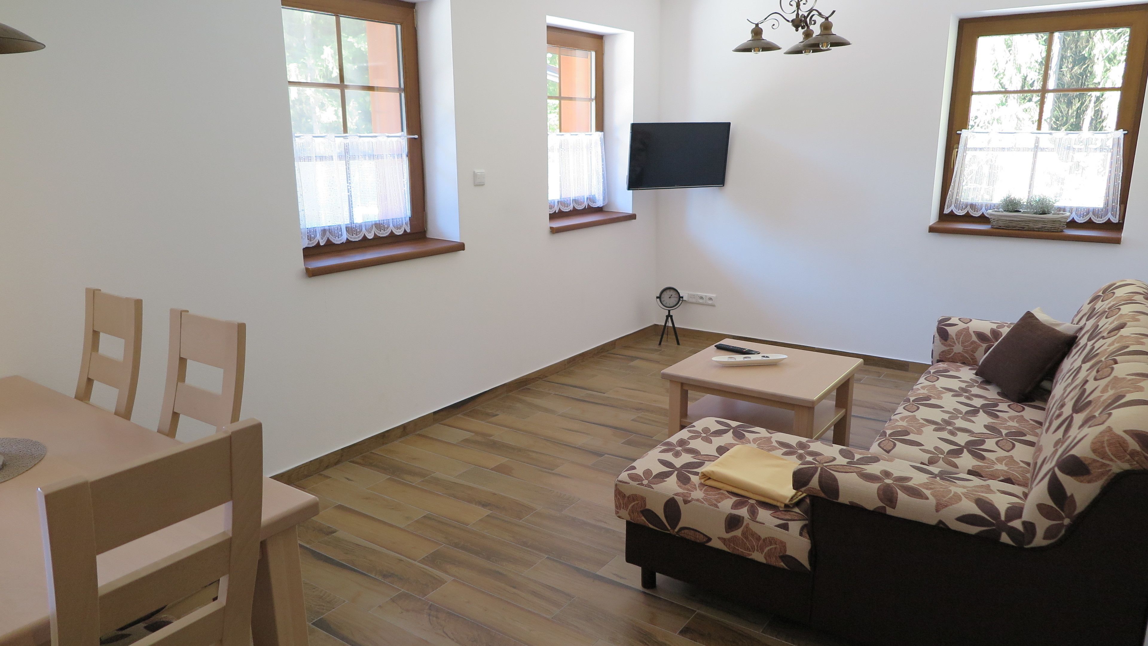 Una sala de estar con un sofá, una mesita y un televisor montado en la pared. Hay ventanas con cortinas y un reloj en la pared.