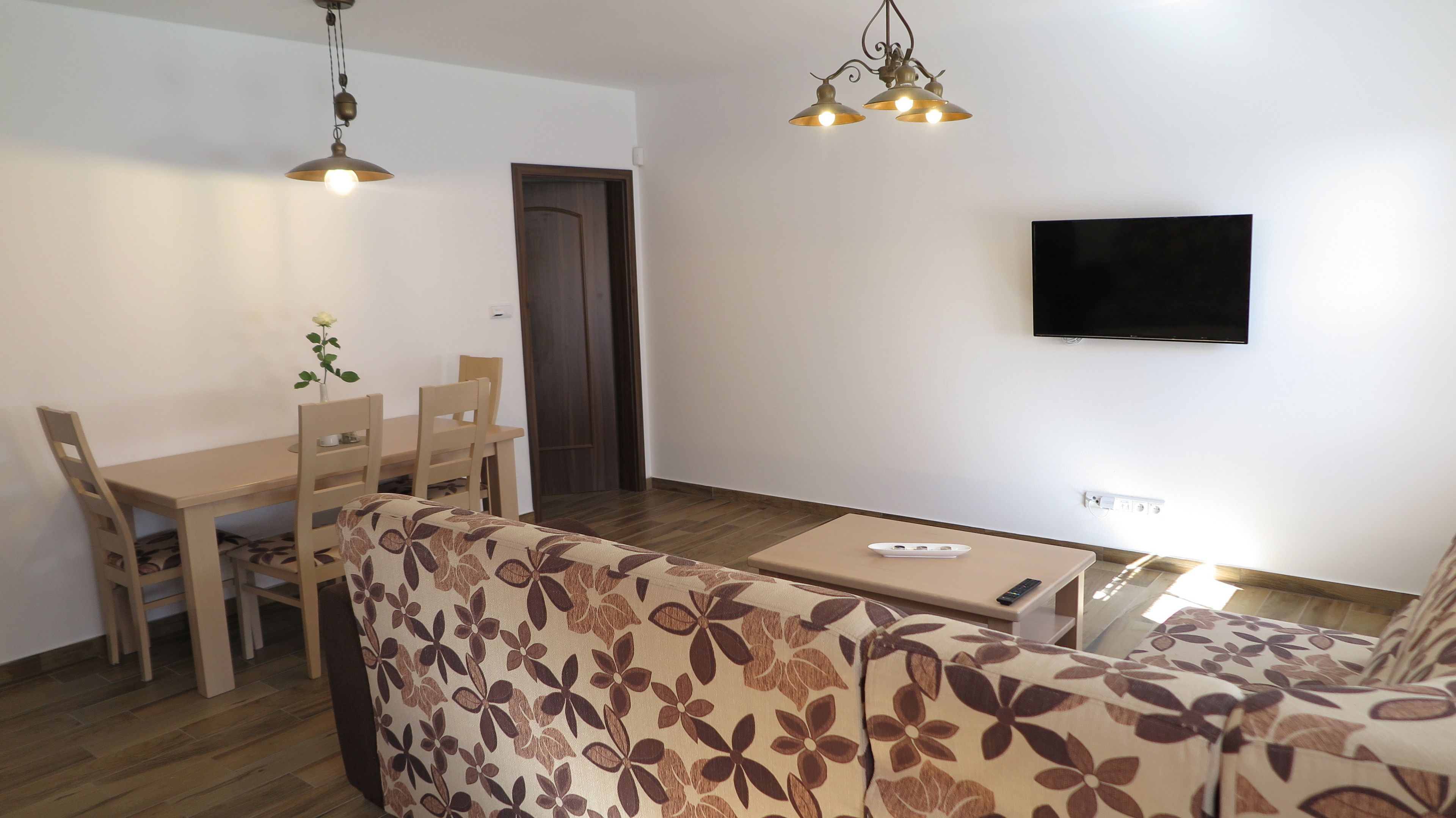 Un saló amb un sofà, una taula de dinar amb cadires, un televisor fixat a la paret i un terra de fusta.