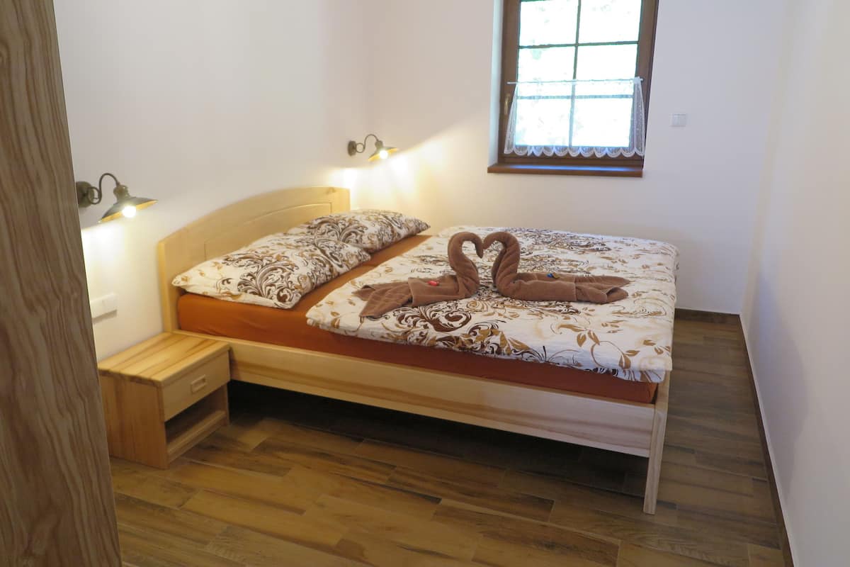 Una cama individual con cabecera de madera y colcha estampada, una mesita de noche pequeña y una ventana con vistas al exterior.