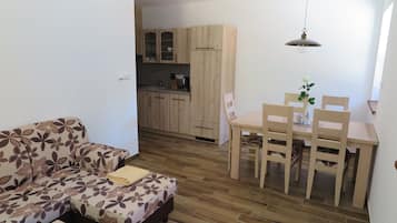 A compact living space with a sofa, a dining table with chairs, and a kitchen area with wooden cabinets and a hanging light fixture.