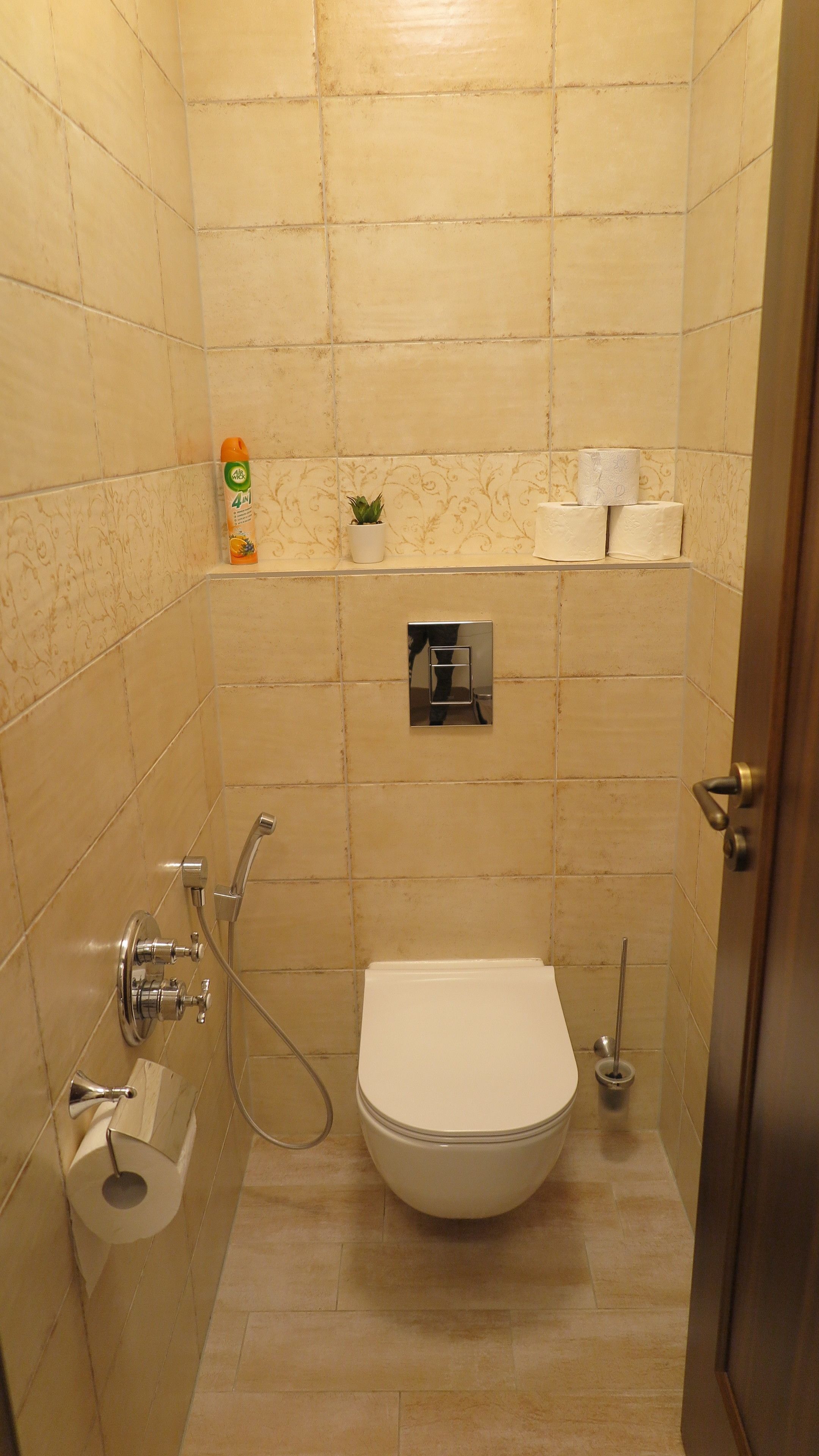 A small, tiled bathroom with a toilet, a hand dryer, a small shelf with a plant and a bottle, and a toilet paper holder.