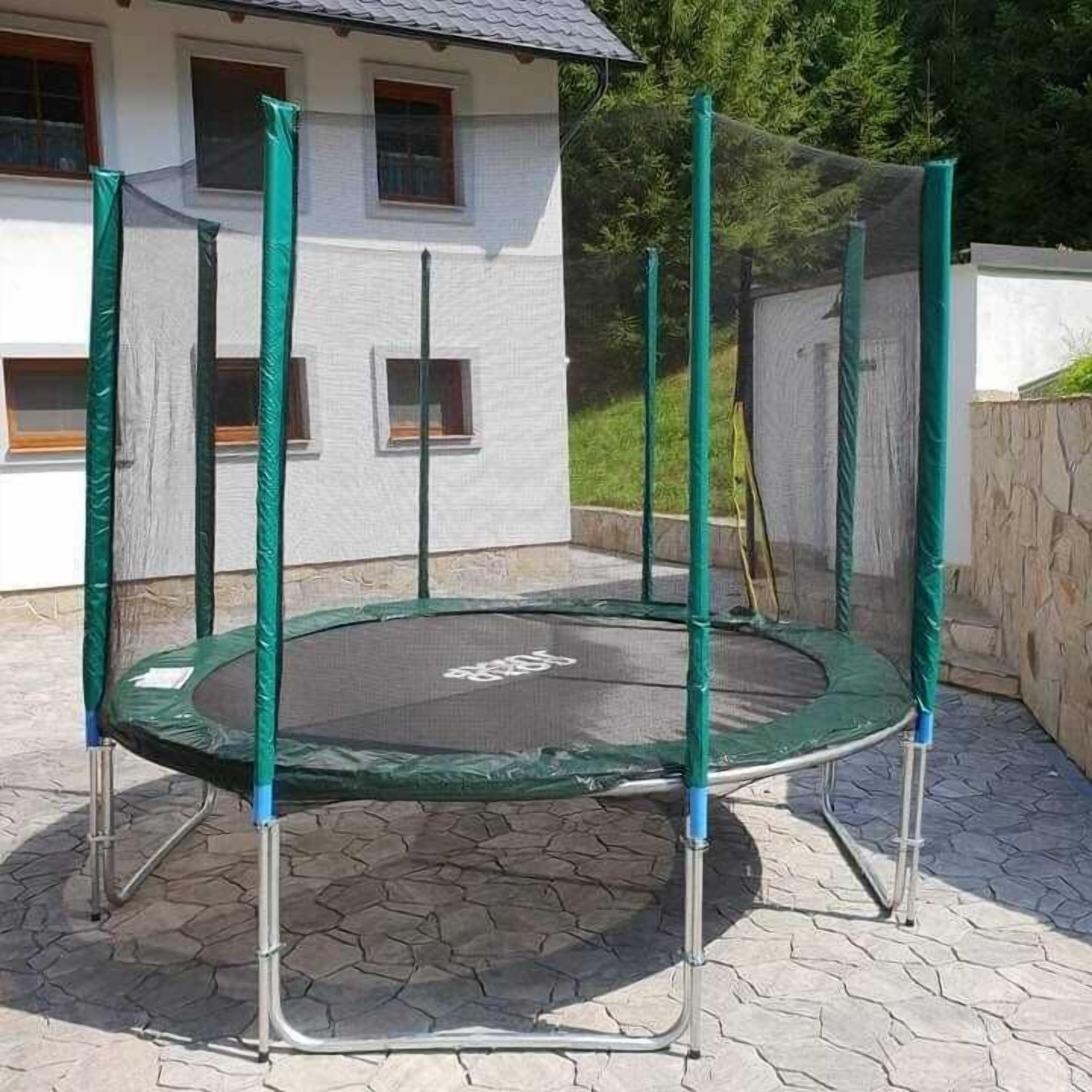 A trampoline with a safety net, set up on a stone patio in front of a white house with a tiled roof.