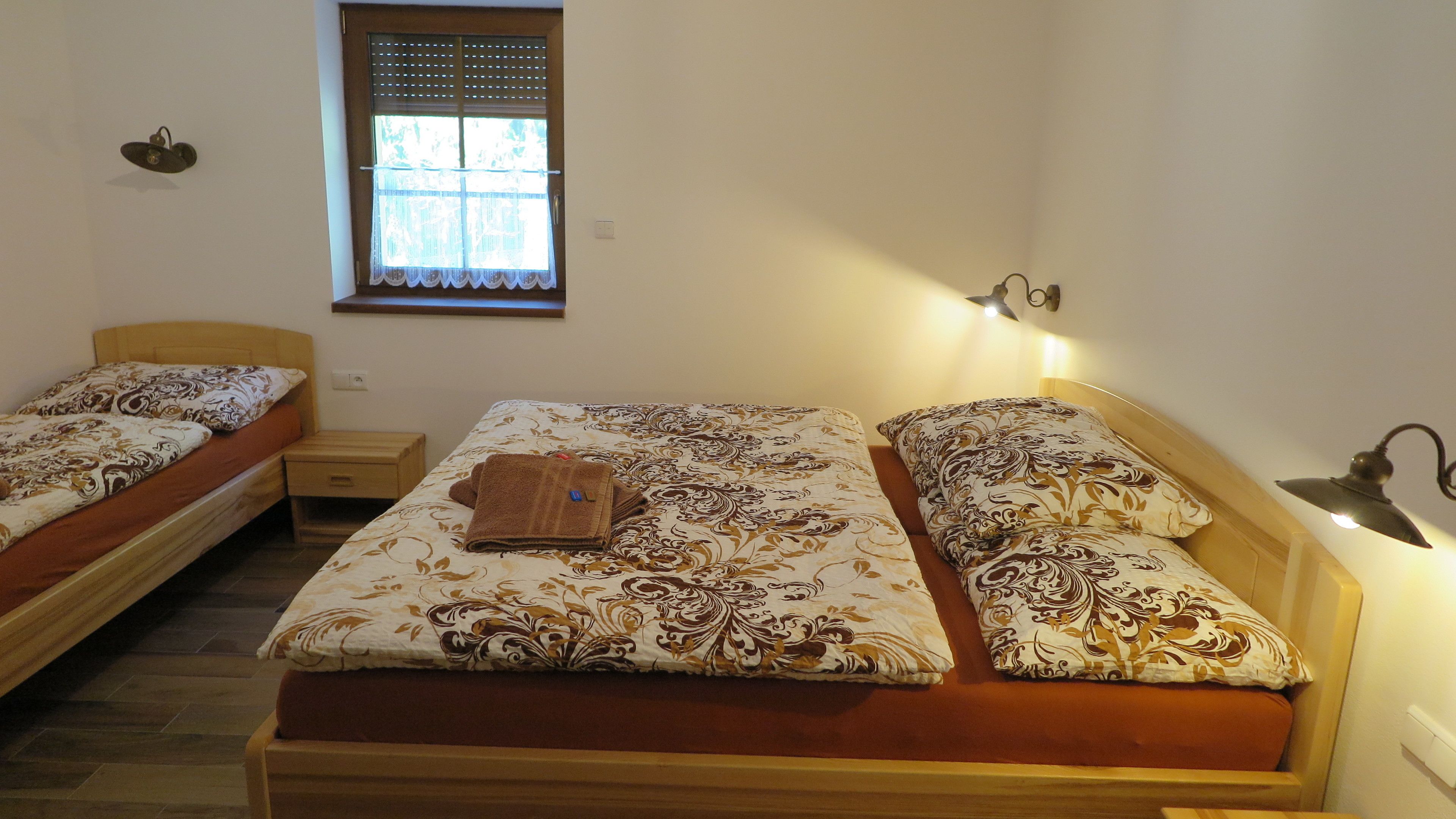 Una habitación con dos camas individuales, una mesita de noche pequeña, una ventana con persianas y un aplique de pared.