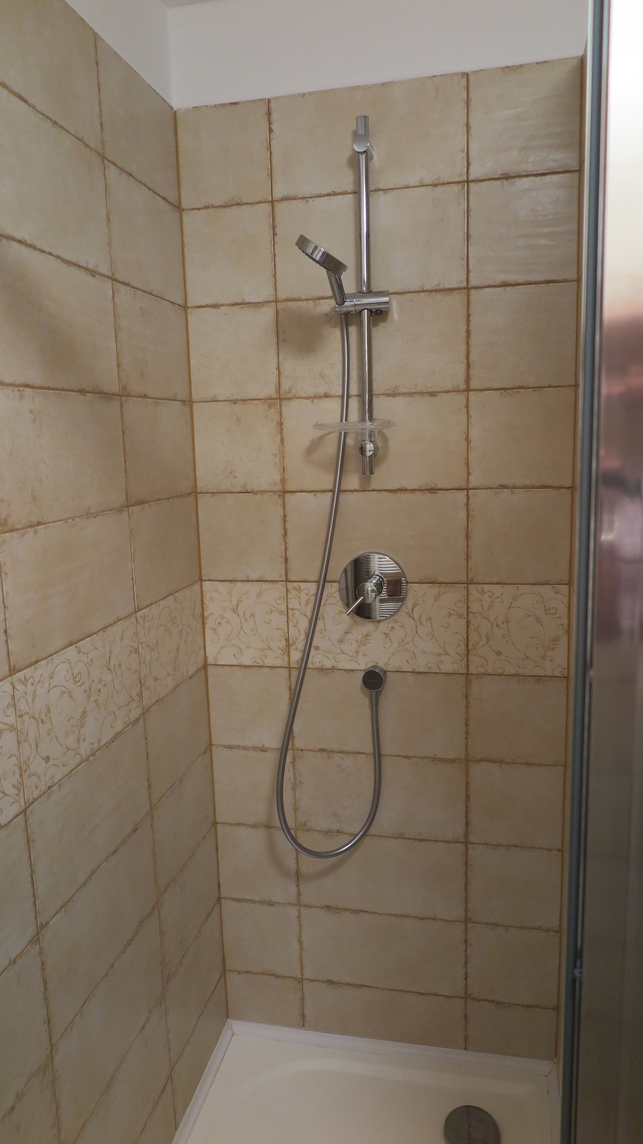 A shower with a handheld showerhead and a wall-mounted faucet, set in a tiled room with a simple design.