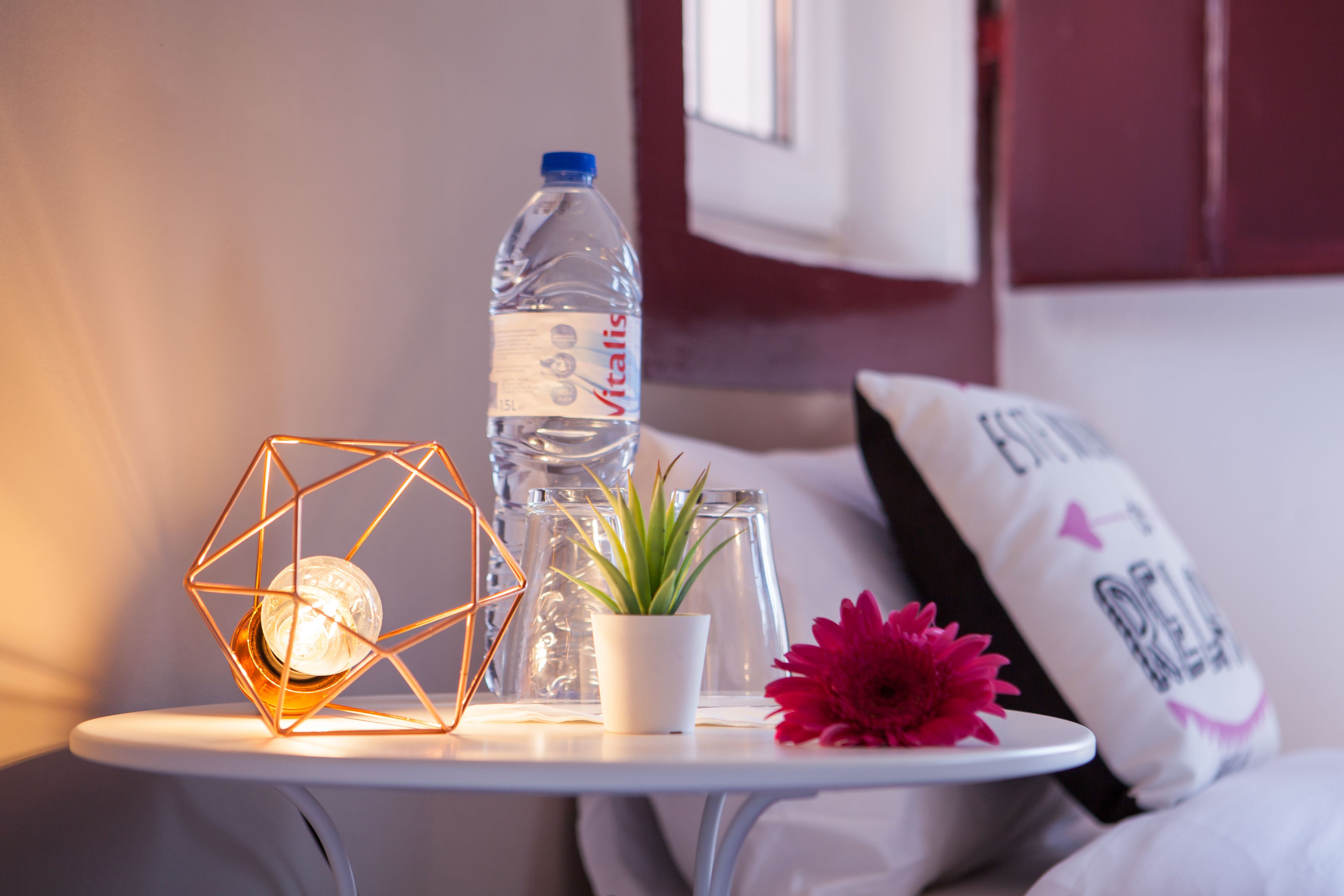 Ein kleiner Tisch mit einer Wasserflasche, einem Blumentopf, einer Blume und einer geometrischen Leuchte.