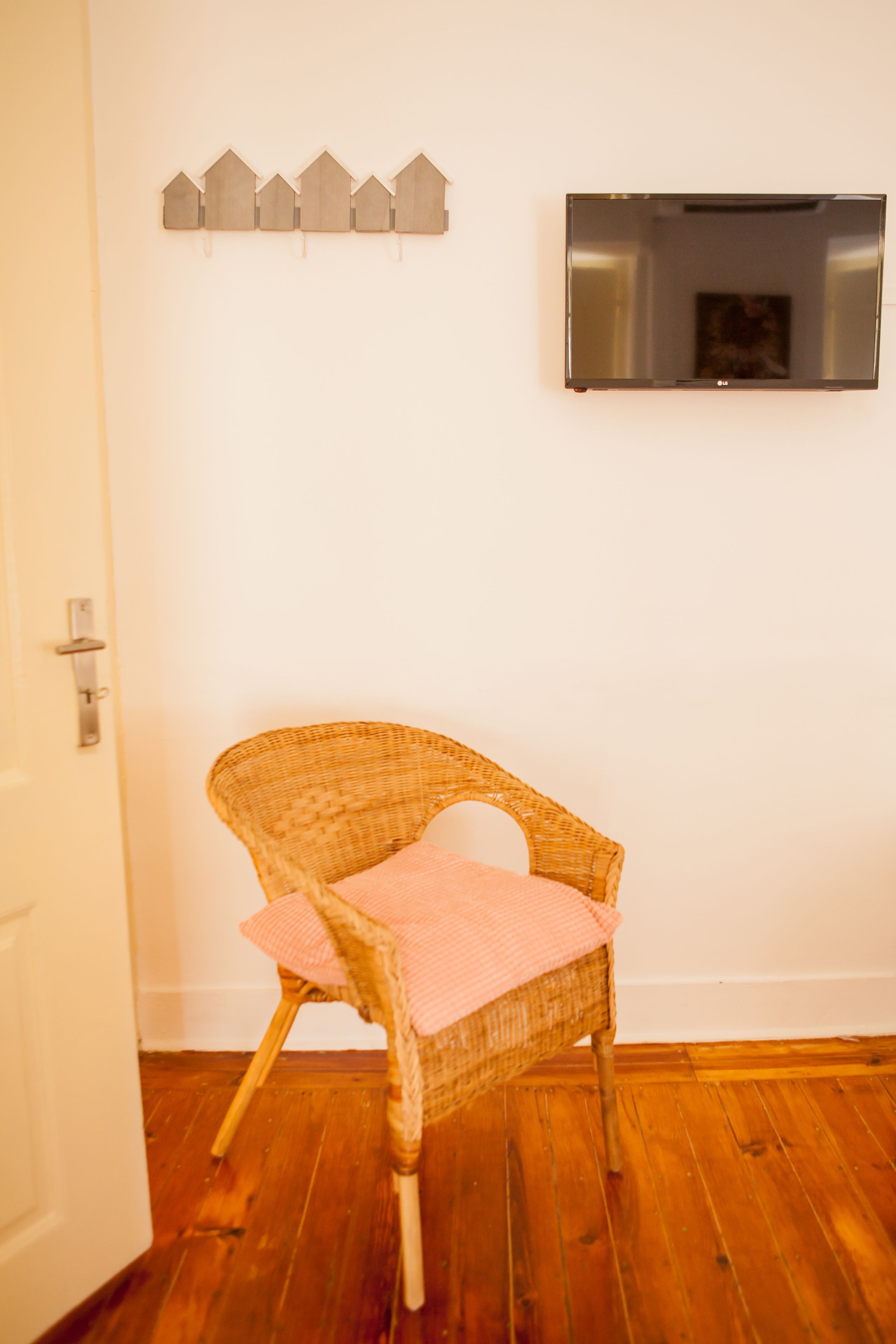 A wicker chair with a pink cushion in a room with wooden flooring, a white door, and a wall-mounted TV above a row of small wooden houses.