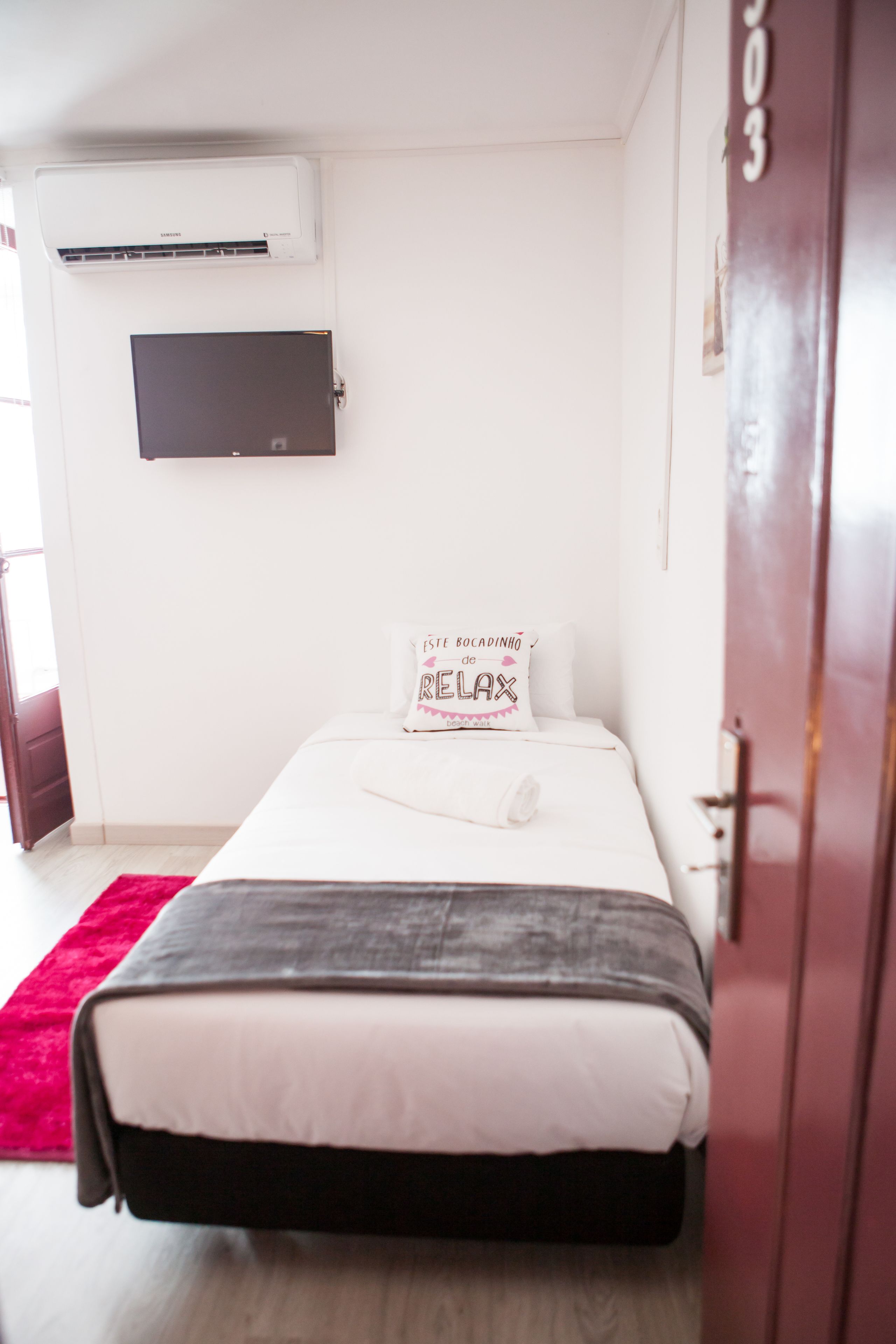 Un dormitorio con una cama, un televisor de pantalla plana en la pared, una puerta con un número y una alfombra roja.