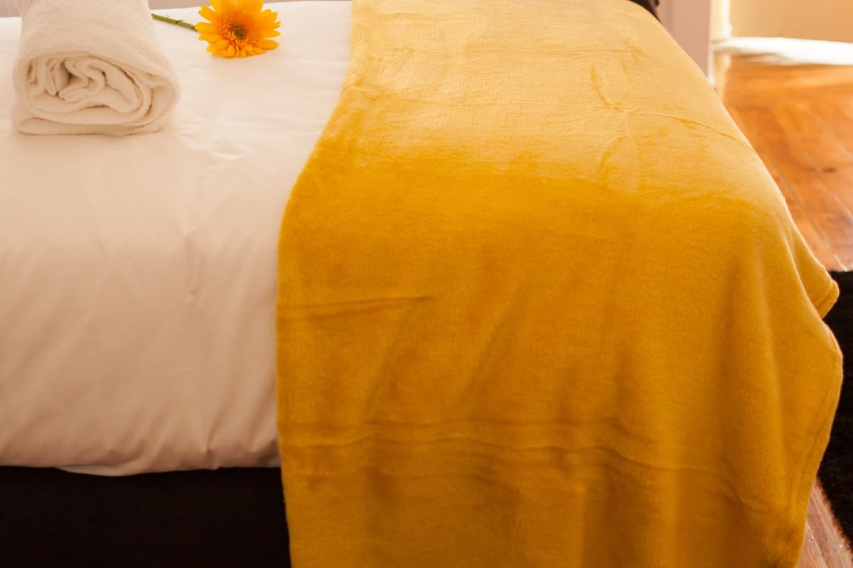 Ett hotellrum med en säng täckt av vita och gula lakan, en ensam gul blomma och ett trägolv.