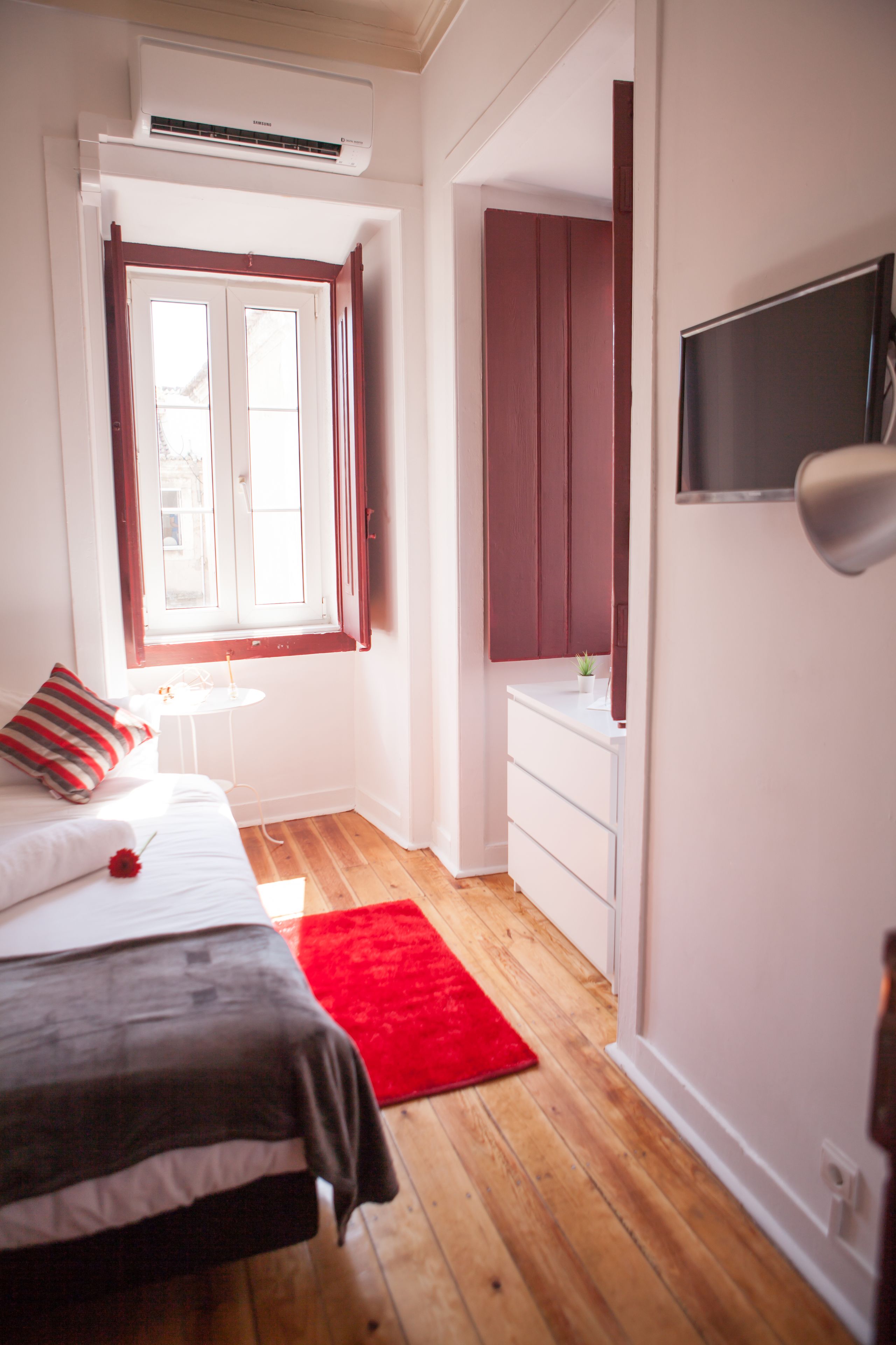 A bedroom with a bed, a red rug, a small table, a TV, and a window with curtains.