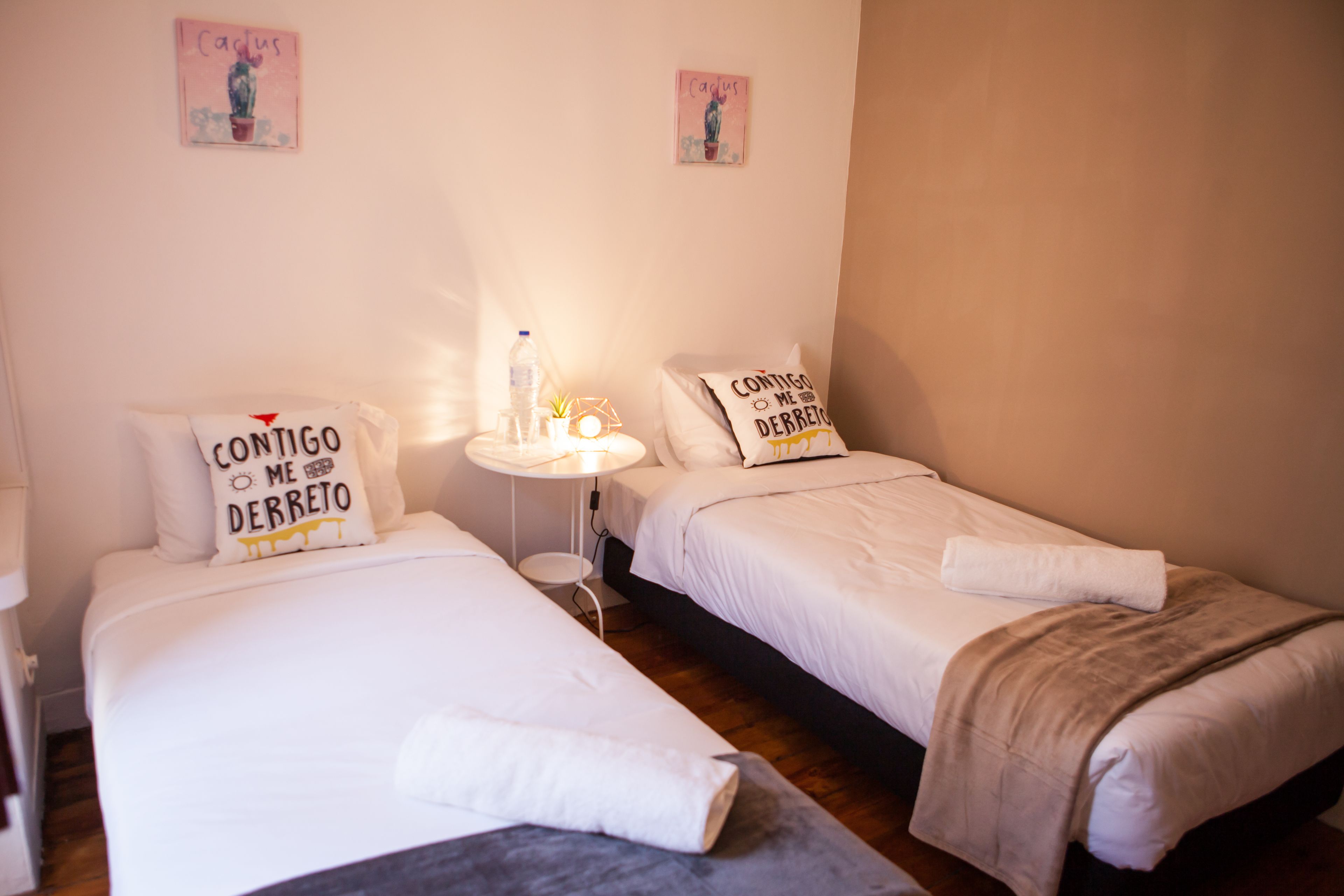 A hotel room with two single beds, each with a pillow featuring a motivational quote. There is a small table with a lamp and a bottle of water.