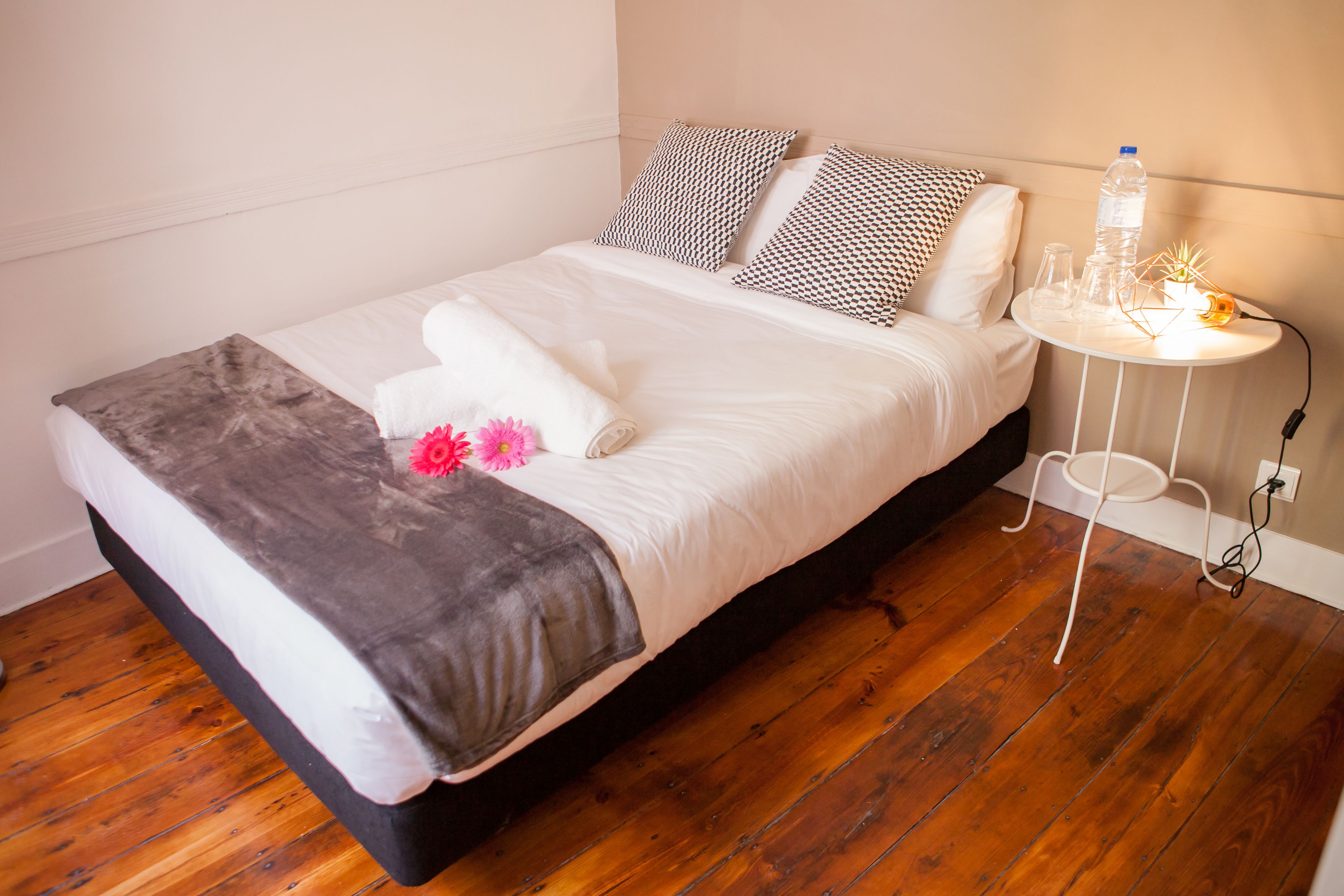 A neatly made bed with a gray and white duvet, a brown throw blanket, and pink flowers. There is a small white side table with a water bottle and a plant.