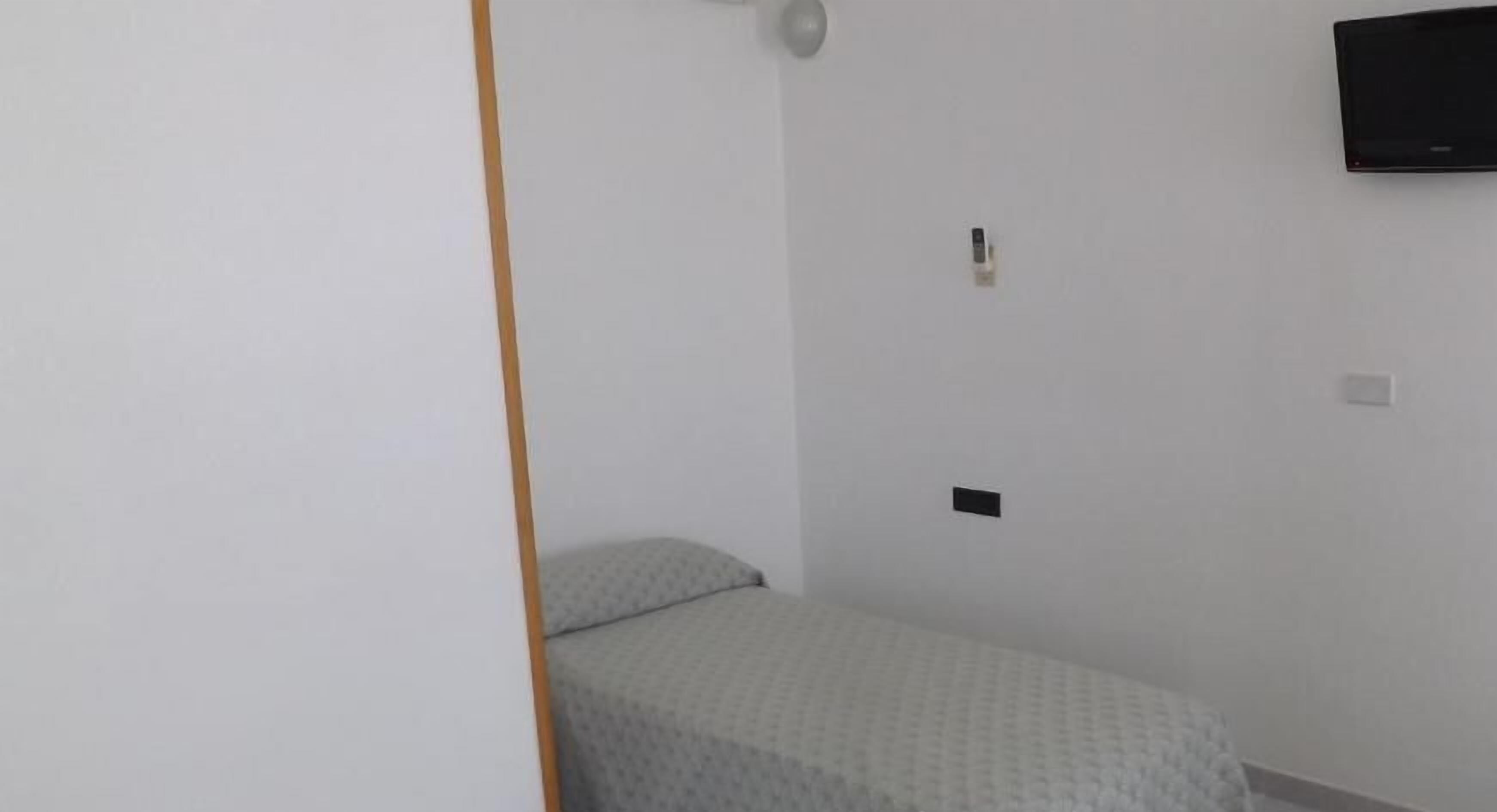 A small, sparsely furnished room with a bed, a wall-mounted TV, and a door.