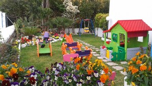 Un jardín con zona de juegos, que incluye tobogán y columpios, rodeado de flores y un invernadero pequeño.