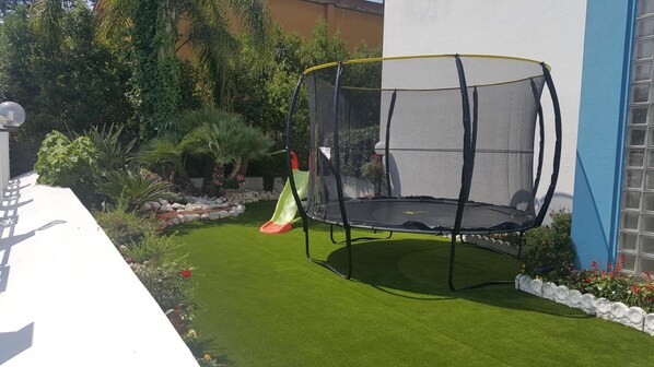 Un trampolín con red de seguridad en un patio trasero con césped artificial, un tobogán y diversas plantas.