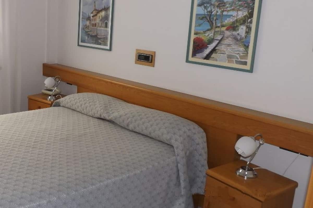 Un lit bien fait, recouvert d’une couvre-lit à motifs, avec une tête de lit en bois, une table de chevet sur laquelle se trouve une lampe, et deux tableaux encadrés accrochés au mur.