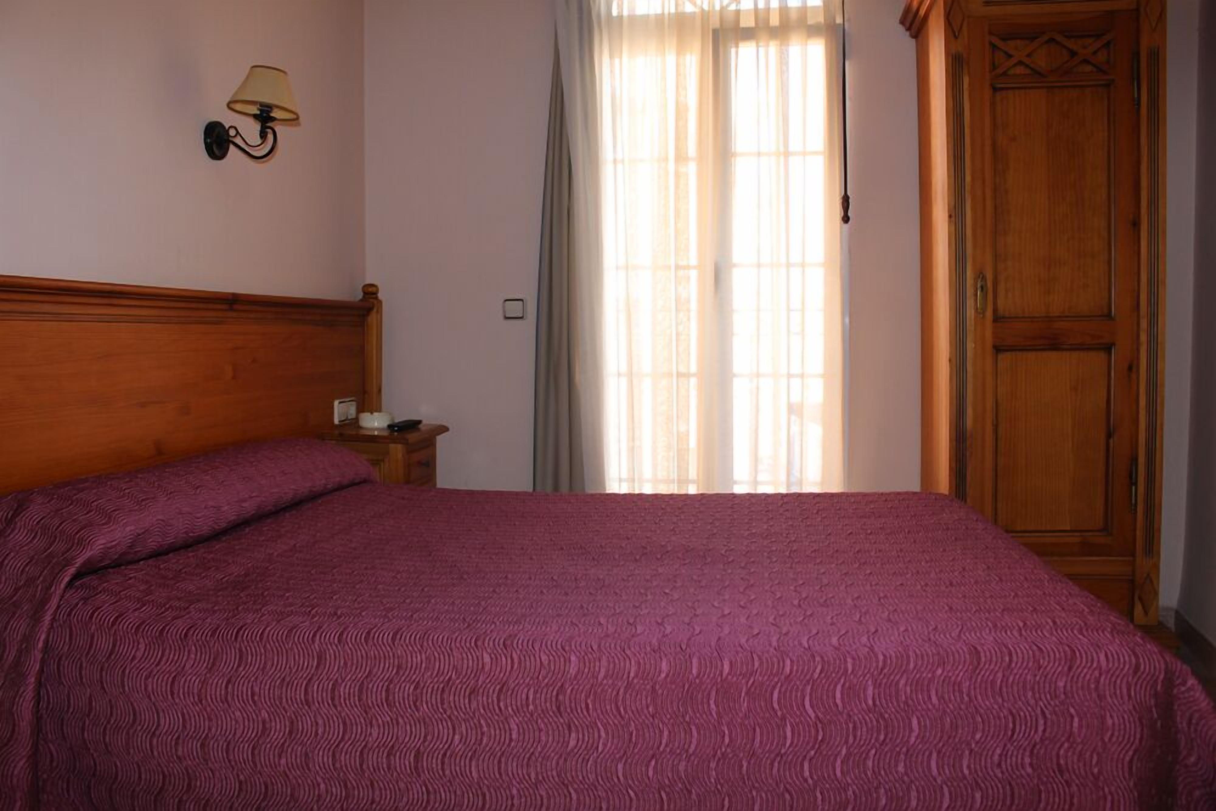 Un dormitorio con cabecera de madera, cama con colcha estampada, mesita de noche con lámpara y armario de madera.